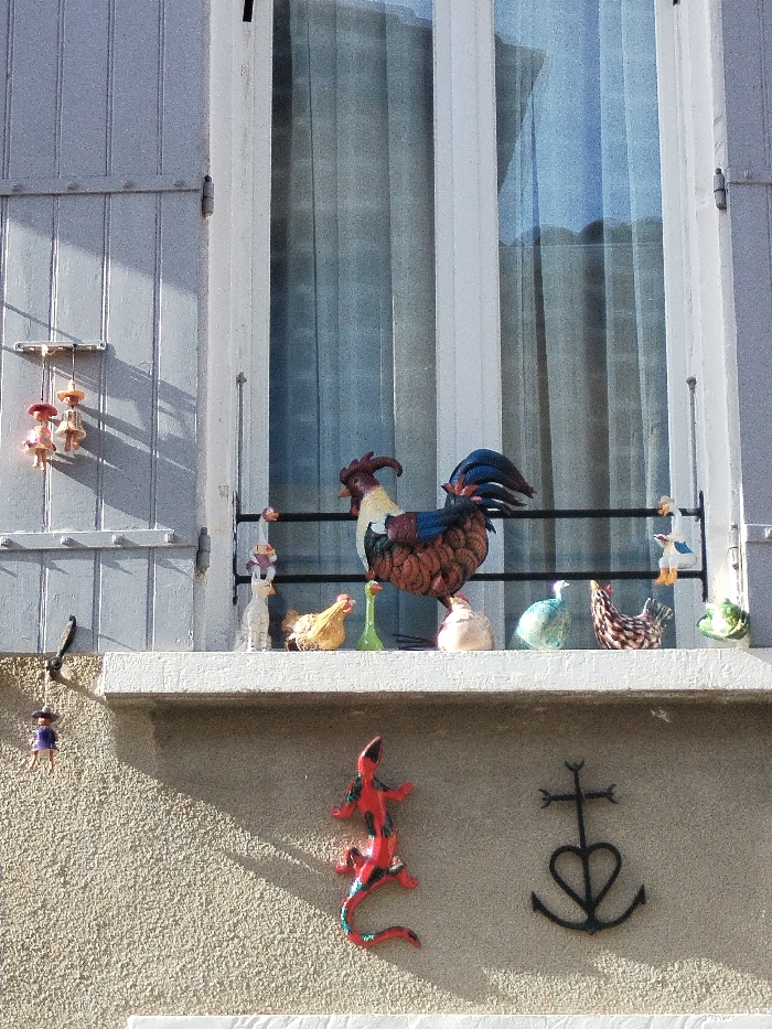 A window with closed grey shutters frames a collection of figurines on the ledge. A colorful rooster dominates, surrounded by various ceramic animals, including chickens and ducks. Below, on the textured wall, a red and black lizard and a dark cross symbol with a heart hang. A small hanging figure dangles near the left shutter, adding a playful touch to the scene. Curtains are visible through the window.