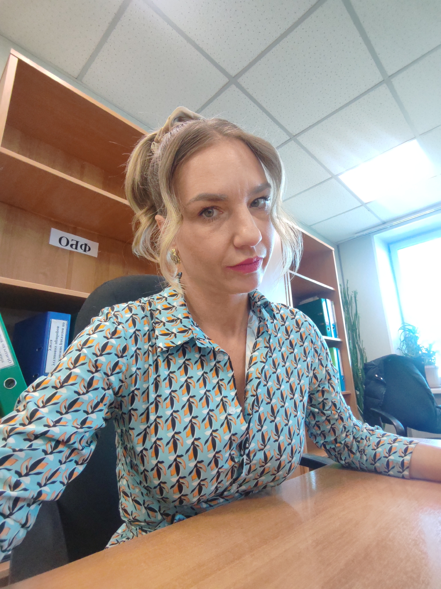The image shows a woman sitting at a desk in an office. She has blonde hair pulled up in a ponytail, fair skin, and is wearing a patterned blue shirt with orange and black leaf-like designs. She is seated in a black office chair, and her hands rest on the light brown wooden desk.
Behind her is a light brown wooden bookshelf containing files and documents. A sign labeled "ОАФ" is visible on the shelf. To the right of the woman, near a bright window, is a coat hanging on the chair. The room appears to be well-lit with fluorescent ceiling lights.
Location: Unknown, Unknown.