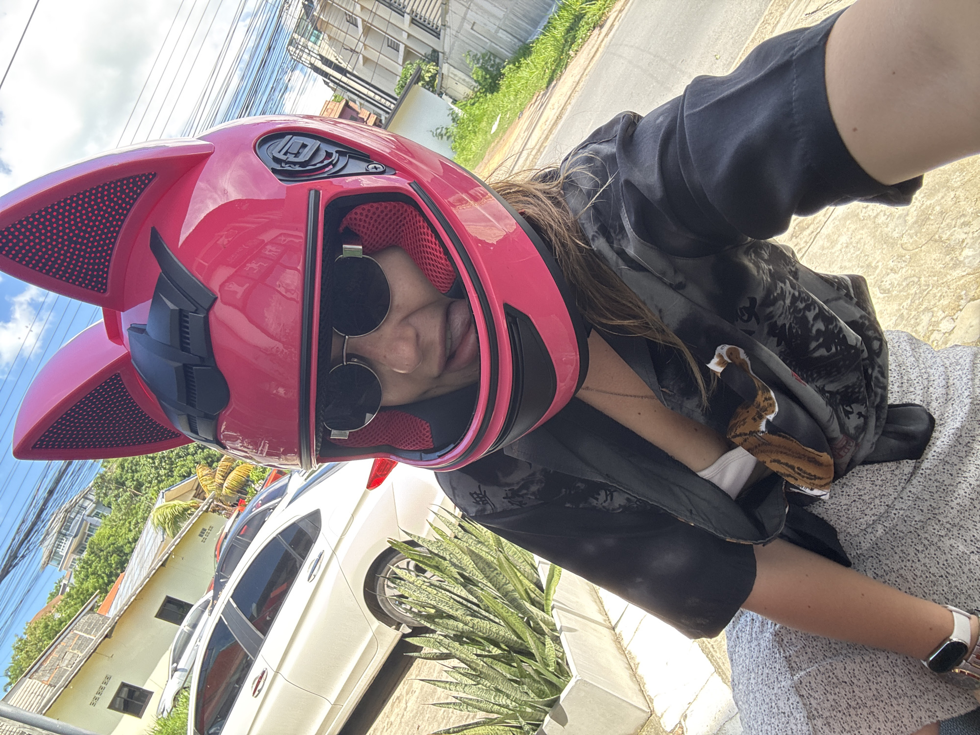 A person wearing a bright pink motorcycle helmet with cat ears and sunglasses is pictured from the chest up, with their tongue sticking out playfully. They are wearing a dark, patterned shirt over a white top and a light-colored skirt. To the left, a white car is parked in front of a building, and a planter with green foliage is visible. The background shows houses, trees, and utility poles under a bright sky, suggesting an outdoor urban or suburban setting in Baan Bang Rak, Thailand.