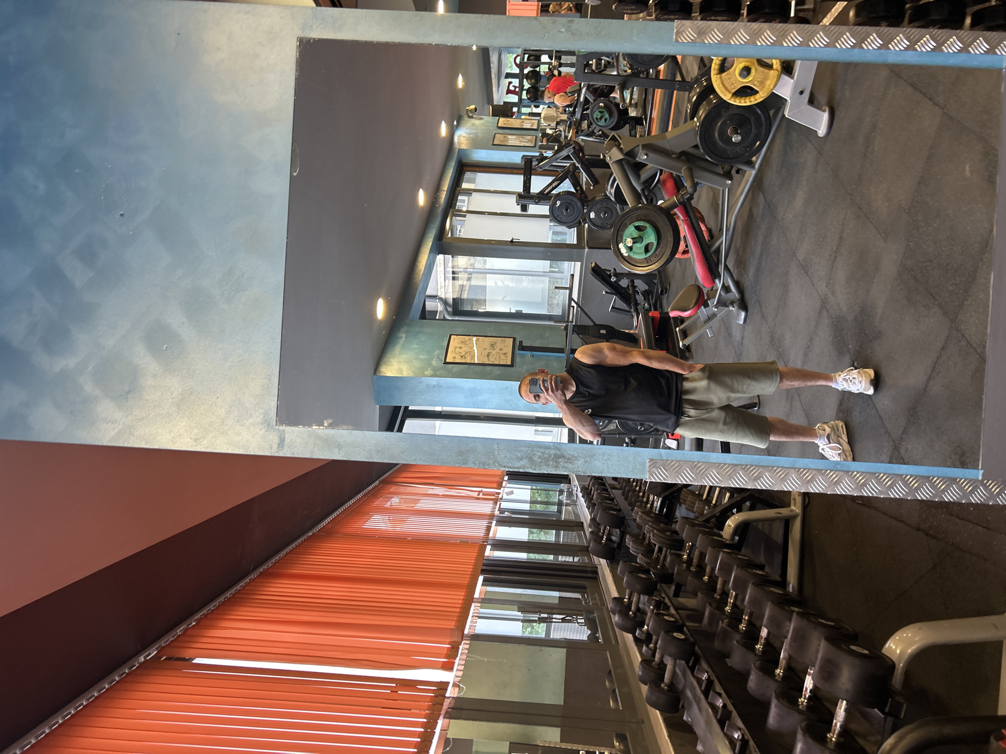 The image captures a selfie taken by a man in a gym, reflected in a large mirror. The man is positioned centrally in the reflection, wearing a black sleeveless top, khaki shorts, and white sneakers. He holds a phone to take the picture, obscuring most of his face, but his short, light-colored hair and muscular left arm are visible.
The setting is a well-equipped gym, likely in Sofia, Bulgaria, as indicated by the context. Various gym equipment is visible in the reflection, including multiple weight machines, barbells, and a long rack of dumbbells in the foreground (bottom of the mirror reflection). Colorful weight plates (yellow, black, green, red) are seen on the machines. The gym floor is covered with dark rubber mats.
The interior features dark walls, some with a textured blue or green finish, and recessed lighting in the ceiling. Large windows in the background allow natural light into the gym, suggesting it is daytime. Outside the windows, some greenery and buildings are faintly visible. On the left side of the image (right side of the actual room, as seen in reflection), bright orange horizontal blinds add a contrasting pop of color. Framed pictures or posters are sparsely placed on the gym walls. In the deeper background reflection, another person wearing a red shirt can be vaguely seen using equipment. No legible text is visible in the image.