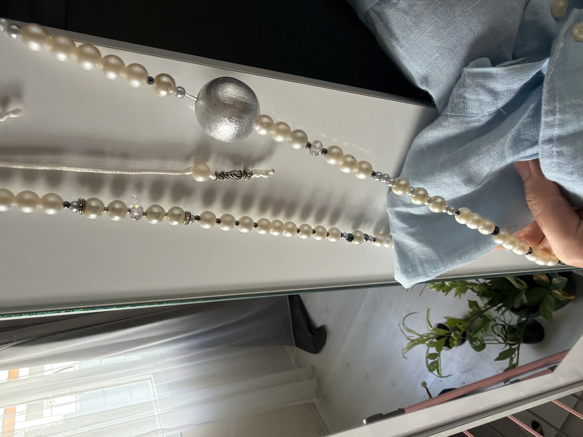 In Izmir, Turkey, a close-up shot shows a pearl necklace with a large, textured silver bead hanging in front of a white surface, likely a cabinet or wall. The necklace is suspended by a thin white string. To the left, a mirror reflects a room with sheer curtains, a window with sunlight streaming through, and several potted plants on a floor. A person's hand, holding a piece of light blue fabric, is visible in the lower right portion of the image. The scene suggests an indoor setting, possibly a bedroom or dressing area.