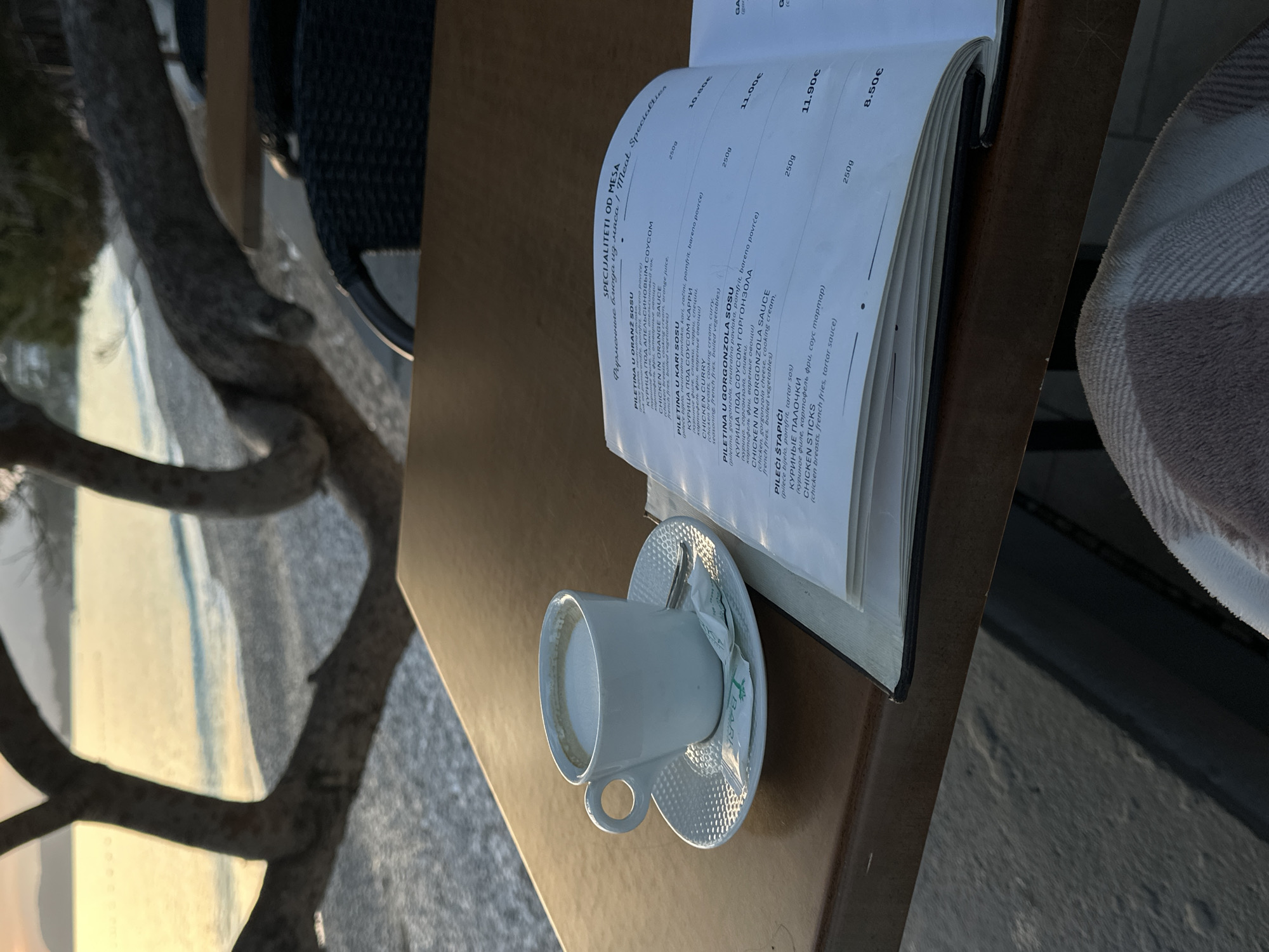 The image shows a table at an outdoor restaurant in Bar, Montenegro. A partially consumed cup of coffee sits on a saucer next to a sugar packet. A menu with Cyrillic and English text listing "SPECIALITETI OD MESA" (meat specialties) leans against the coffee cup. The table has a brown, wood-like finish. A blurred beach scene is visible in the background, suggesting a seaside location. A tree branch is also visible on the left of the image. A chair is partially visible. A blanket is on another chair nearby.