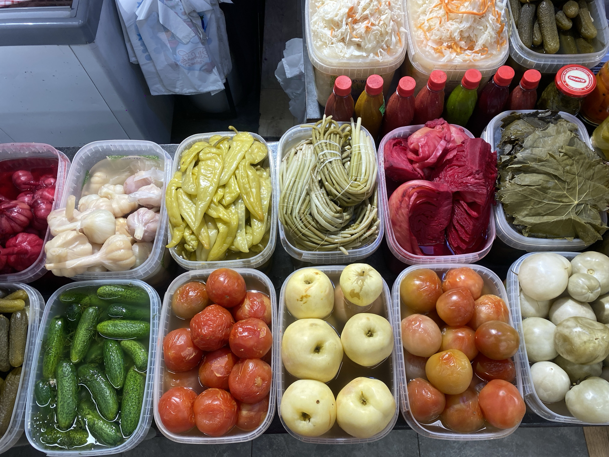 This overhead shot captures a vibrant display of various pickled and fermented goods, likely at a market stall in Moscow, Russia. The scene is dominated by numerous transparent plastic containers and a few glass jars, all filled with different foods, neatly arranged across a dark surface.
In the top row, large containers hold white, shredded sauerkraut-like fermented cabbage, some mixed with carrots, alongside containers of small dill pickles. Behind these, a line of small bottles with red and green caps, likely various sauces or dressings, are visible.
The middle row showcases a diverse array of pickles. From left to right, there are red pickled garlic bulbs in a reddish brine, whole pickled garlic cloves, bright yellow pickled peppers, and bundles of long, slender pickled garlic scapes tied with white string. Next to these are striking red pickled cabbage pieces and containers filled with dark green pickled grape leaves.
The bottom row features more pickled vegetables and fruits. There are small green pickled cucumbers, followed by containers of whole pickled red tomatoes and pale yellow pickled apples. The final container on the right holds what appear to be pickled green or white tomatoes, or similar small, round fruits.
One notable detail is a red lid on a glass jar in the upper right quadrant, clearly showing the Cyrillic text "ПИКАНТА" (PIKANTA), a brand name that aligns with the specified location context of Russia. The overall impression is one of abundance and variety, characteristic of a food market specializing in traditional preserves. The bright, even lighting suggests an indoor setting.