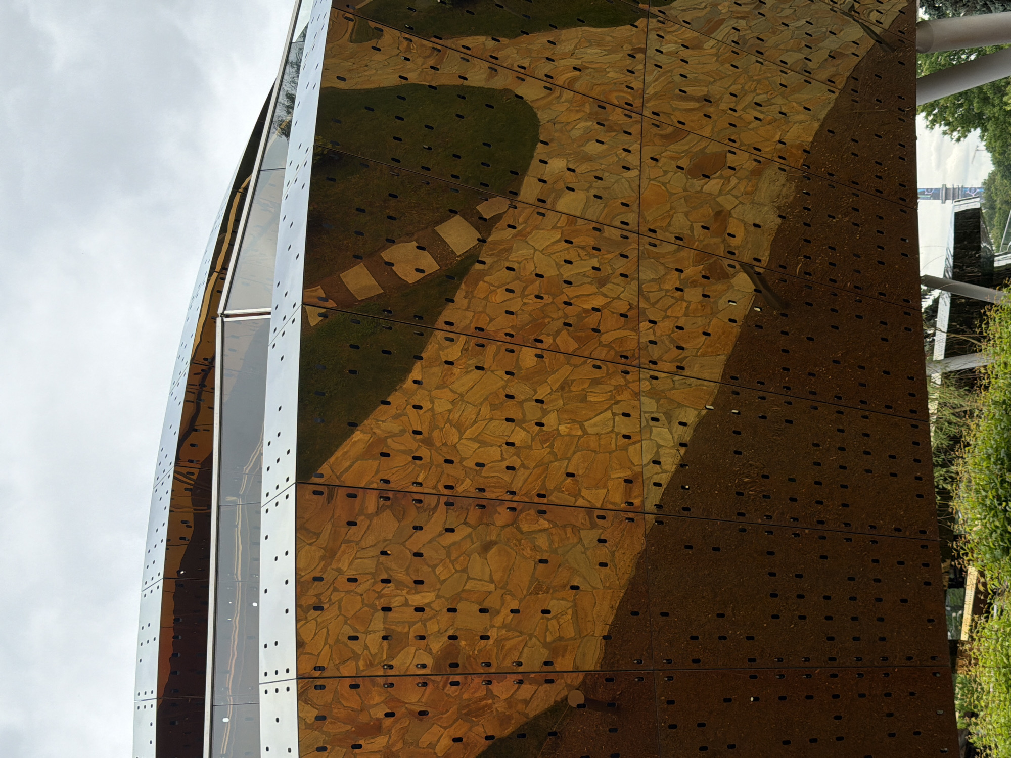 The image presents a close-up, rotated view of a modern building facade under an overcast sky during the daytime. The building's exterior is constructed from numerous rectangular panels with a highly reflective, bronze or golden-toned surface. Each panel is uniformly perforated with rows of small, elongated holes.
The reflective quality of the facade creates a distorted and abstract mirror image of the surrounding environment. Prominently reflected across much of the building's surface are elements of a landscaped area. These include vibrant green foliage, indicative of trees or bushes, particularly visible on the right side of the image. Further to the left within the reflections, a patterned ground surface is discernible, featuring a pathway composed of irregularly shaped, light-colored stones or pavers interspersed with patches of green grass.
The building exhibits a dynamic, possibly faceted or curvilinear architectural design, where sections of the facade appear to curve or angle differently, creating varied reflections. The top left portion of the image shows a direct view of the bright, cloudy sky. In the far right reflection, hints of other structures, such as white pipes or beams, are faintly visible. There are no people or discernible text in the image. The scene emphasizes the interplay between modern architecture and its natural surroundings through the medium of reflection.