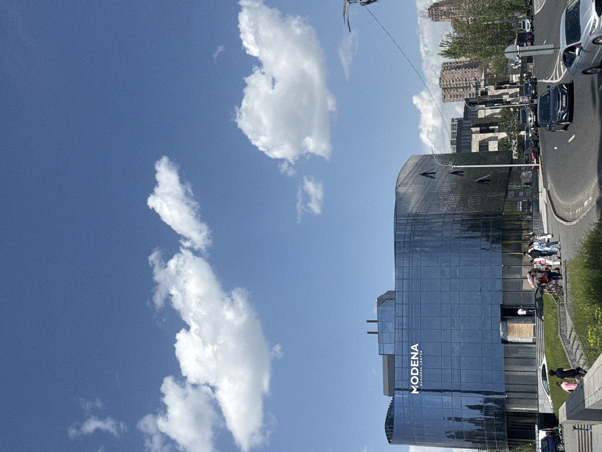The image shows the Modena Business Center, a modern glass building, in Yerevan, Armenia. The sky is blue with scattered white clouds. The building's facade reflects the sky. Adjacent to the building, there is a road with cars and a sidewalk with people walking and standing. In the background, there are residential buildings and trees.