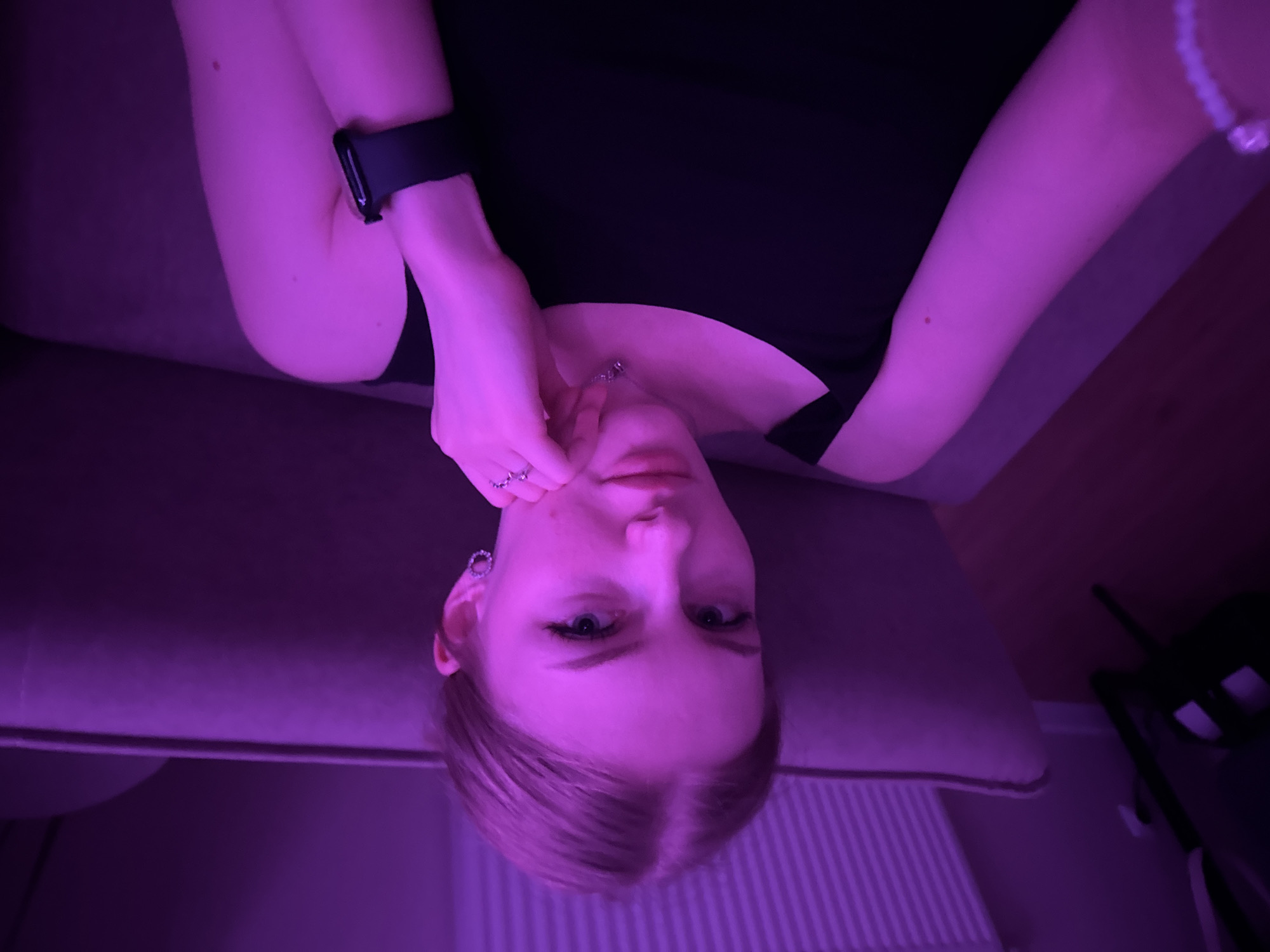 A woman is lying on a purple couch. She has fair skin, short blonde hair, and is wearing a black top. She is holding her face with one hand, wearing a ring on one finger. There appears to be a watch on her wrist. The environment is dimly lit with purple-toned lighting. A radiator is partially visible in the background. The city of Perm, Russia is not identifiable from this image.