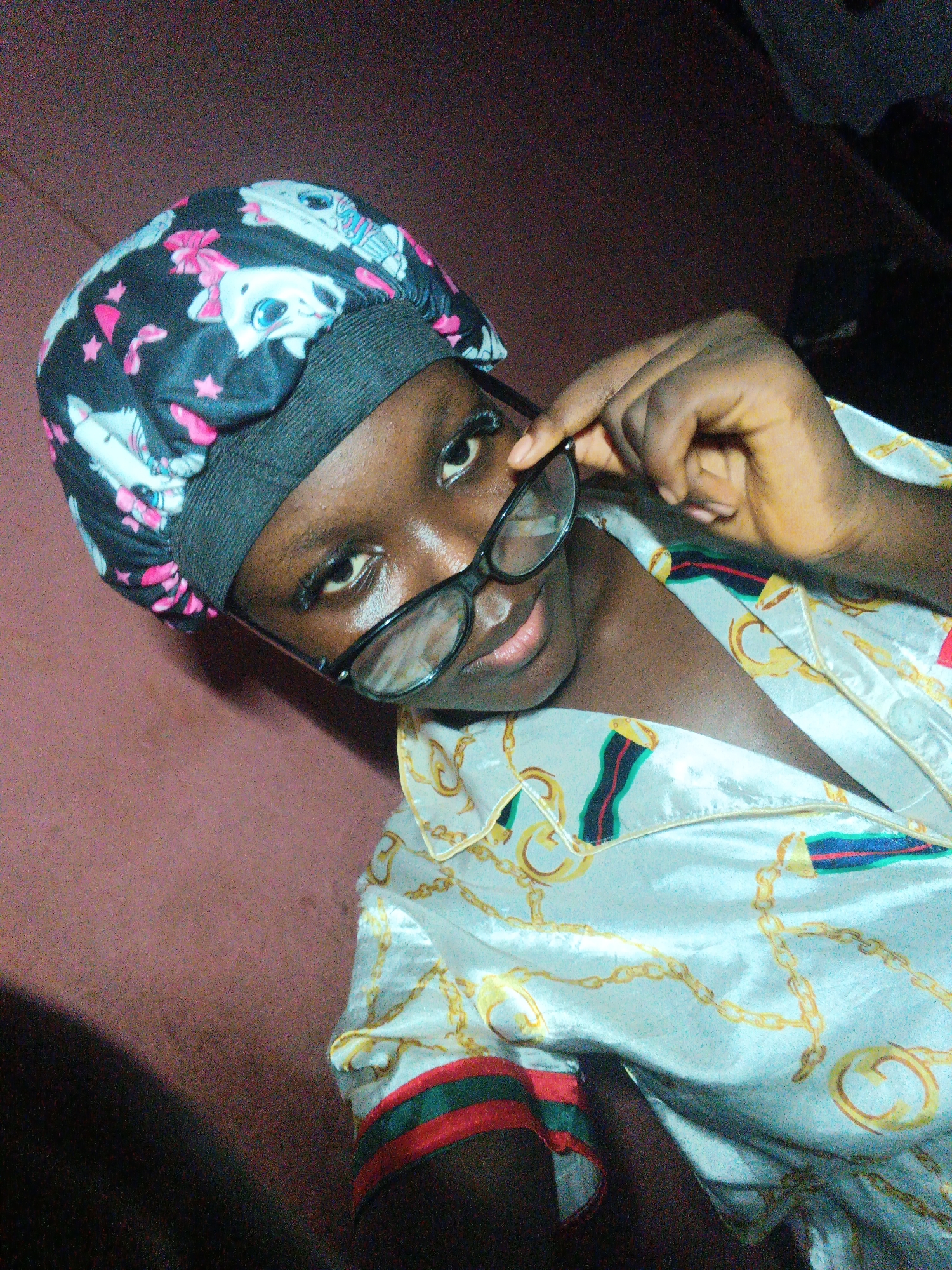 The image captures a close-up portrait of a young person, likely in an indoor setting in Onitsha, Nigeria. The subject, with dark skin, looks directly at the viewer with a confident and slightly playful expression, peering over dark-rimmed glasses held by their right hand (viewer's left).
The person wears a black head covering, possibly a bonnet or satin cap, which is patterned with white cartoon cat faces featuring large blue eyes and pink bows, alongside pink stars. A dark elastic band secures the headwear. Their attire includes a light-colored, likely satin or silk-like, shirt. The shirt is adorned with an elaborate pattern of golden chains and ornate rings, and its collar and cuffs (partially visible) feature a distinct striped design in red, dark green/blue, and white.
The background consists of a plain, reddish-brown wall or surface, appearing somewhat grainy. A darker, indistinct area is visible in the upper right quadrant of the frame. The lighting seems artificial, possibly from a flash, creating highlights on the subject's skin and the glossy fabric of the shirt. No discernible text is visible within the image.