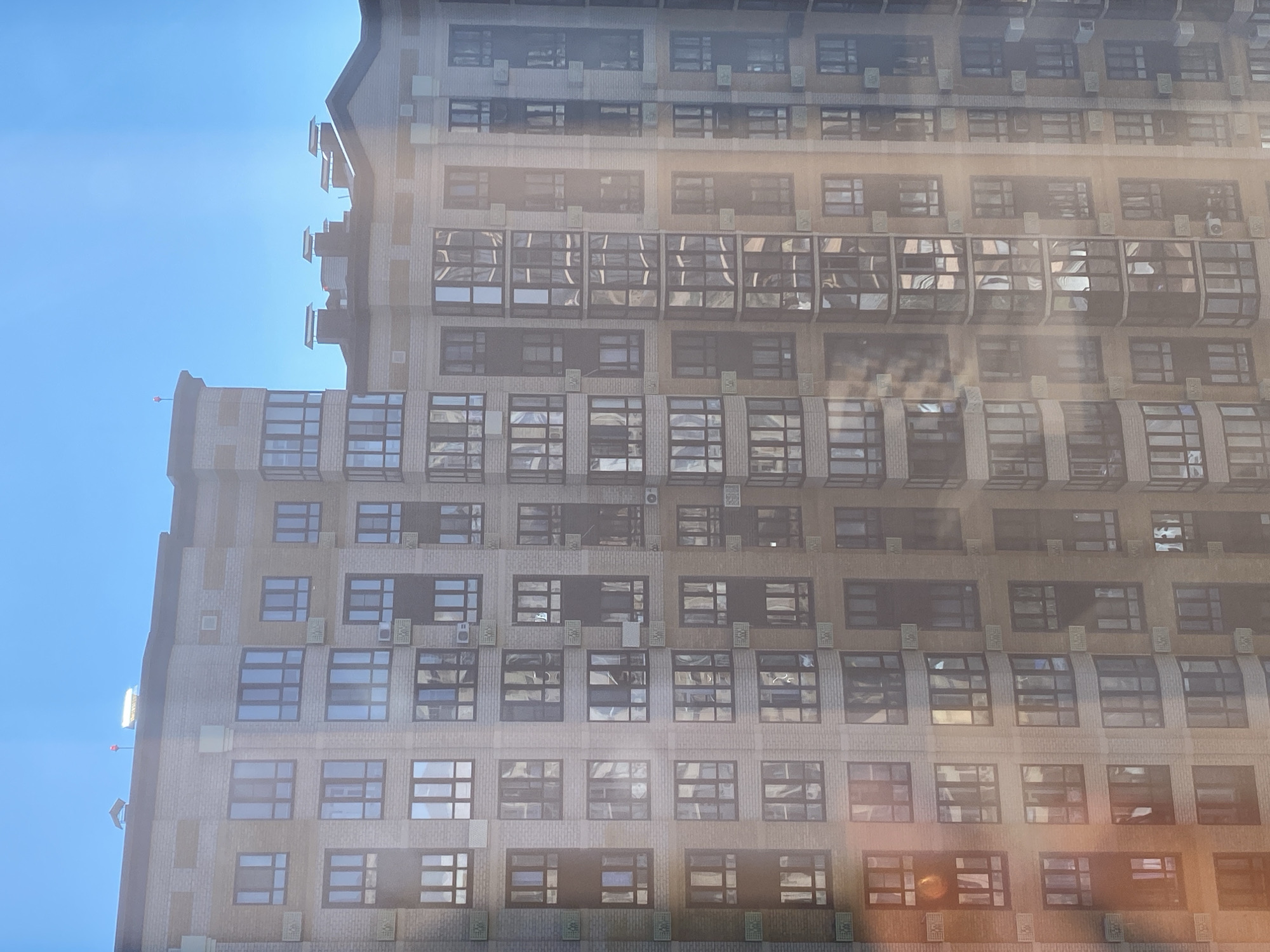 A high-angle, medium shot captures a sunlit, modern apartment building under a clear blue sky. The building, constructed with light-colored brick and accented with darker brown bands, features numerous windows arranged in a grid pattern. Many windows are dark, reflecting the sky and surrounding structures, while others appear lighter, suggesting interiors or different lighting conditions. Air conditioning units are visible mounted on some exterior walls. Several chimneys or vents rise from the rooftop, and a red blinking light is present on a raised structure on the left side of the roof. The building appears to be tall and multi-story, with some sections projecting outward, creating a complex facade. The image is slightly hazy, possibly due to sunlight or atmospheric conditions, creating lens flares and a soft glow over the scene. There are no people visible in the image. The overall impression is that of a contemporary urban residential environment bathed in daylight.