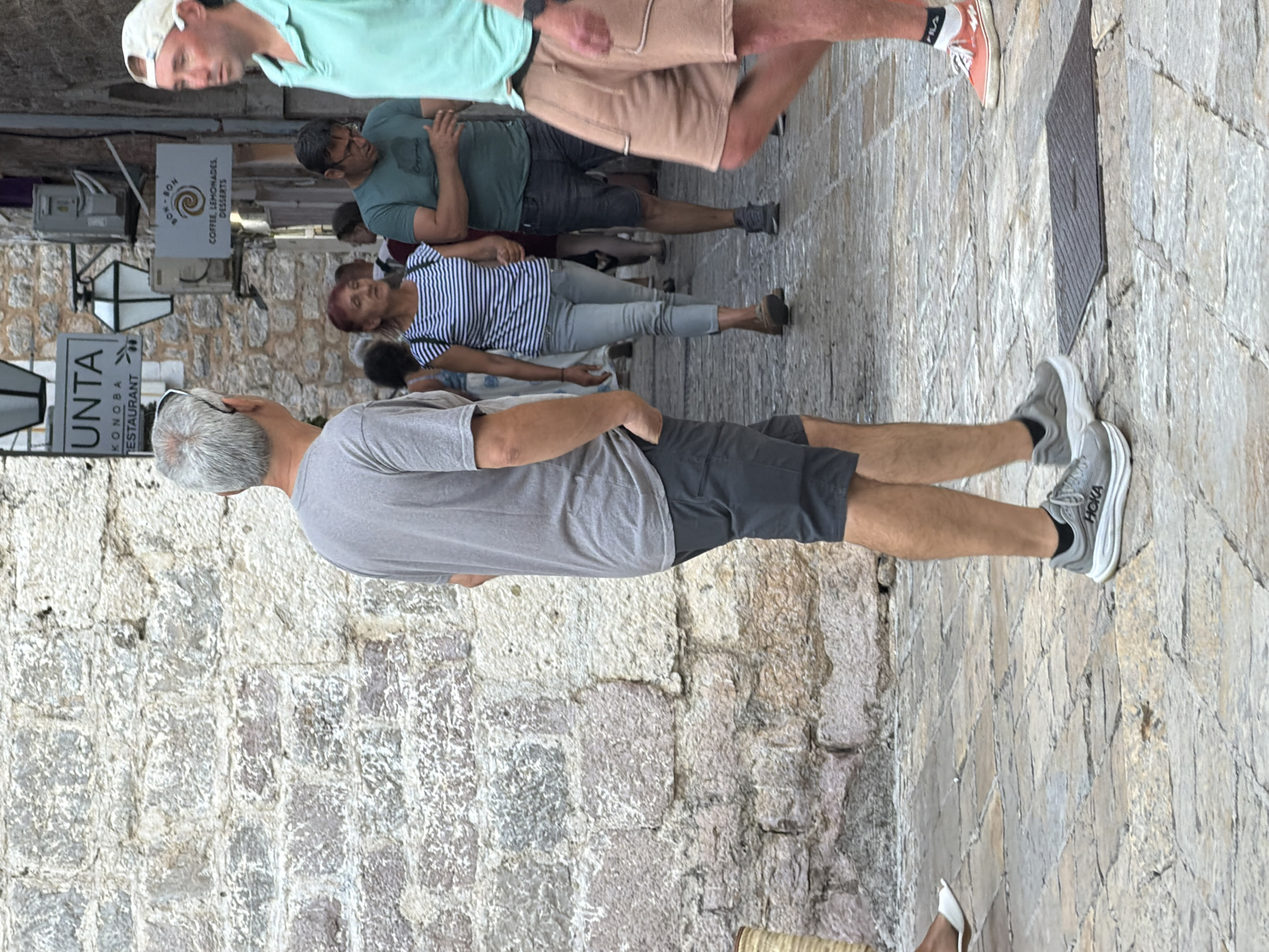 The image captures a street scene in the old town of Budva, Montenegro. A man with graying hair, wearing a grey t-shirt and dark shorts, is walking away from the camera. To his right, a woman with red hair, wearing a striped top and light-colored pants, is walking towards the camera. Behind her, a man in a green t-shirt and dark shorts is also walking. Further to the right, another man in a light green polo shirt and tan shorts is walking away from the camera. The street is paved with cobblestones, and the buildings are made of stone. Signs for "UNTA KON OBA RESTAURANT" and "BON BON COFFEE, LEMONADES, DESSERTS" are visible on the buildings.