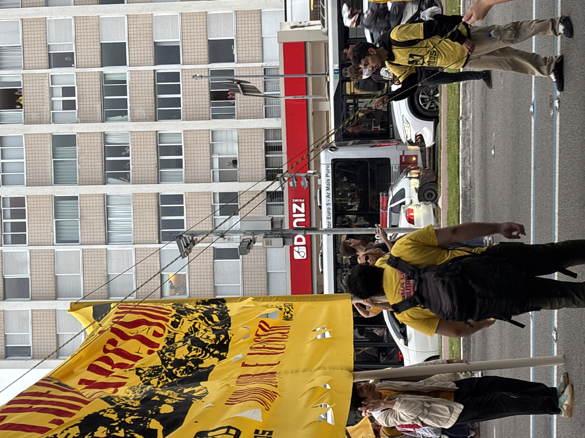 A vertical eye-level outdoor shot captures a public demonstration in São Paulo, Brazil. A large, yellow, tattered banner with black and red markings is prominently displayed, obscuring the lower portion of the frame. Behind the banner, a multi-story building with numerous rectangular windows is visible. The building appears to be made of a light-colored material. A pole is seen extending upwards, with electrical wires running diagonally across the frame. To the right of the building, a Diniz Prime sign is visible. A bus and several cars are parked on the street. People are marching along the road, some wearing yellow shirts. The road is paved with markings, suggesting it's a city street. The environment is typical of an urban setting with buildings, transportation, and public gathering.