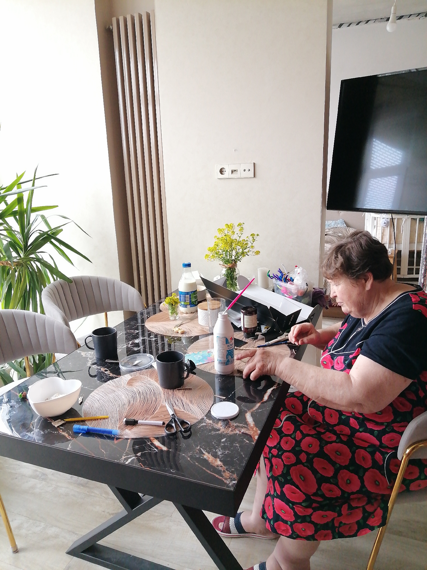 An elderly woman sits at a black marble-patterned table in a room. She wears a black dress with red poppy patterns and sits on a grey chair with gold legs. The table is cluttered with crafting supplies, including a white bowl, pens, scissors, bottles, mugs, and placemats with circular designs. A vase of yellow flowers and a houseplant sit on the table near a window with a beige radiator. A flat-screen television is mounted on the wall. The image was likely taken indoors, possibly in Trubacheyevka, Russia. The woman appears to be engaged in a crafting activity.