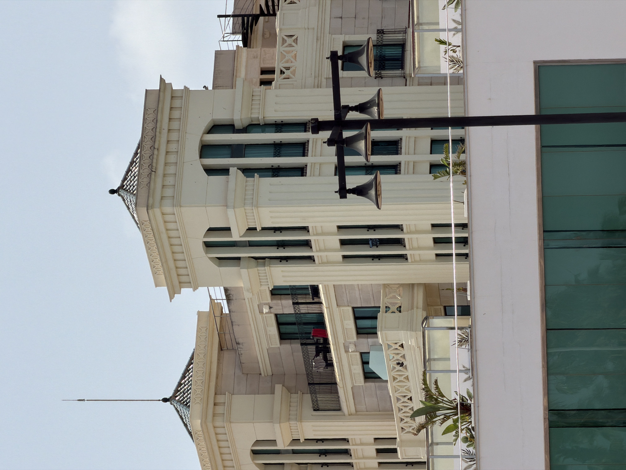 The photograph captures a modern apartment building exterior under a pale sky. The building features a beige facade with ornate architectural details, including fluted columns and decorative moldings around the windows and balconies. Several balconies are visible, some with black wrought-iron railings, furnished with outdoor seating and potted plants. One balcony has a red object on a chair. A prominent black lamppost with four downward-facing lamps stands in the foreground. In the background, a spiral staircase is visible on another section of the building. The lower part of the image shows a white wall topped with green glass panels. The scene is devoid of people.