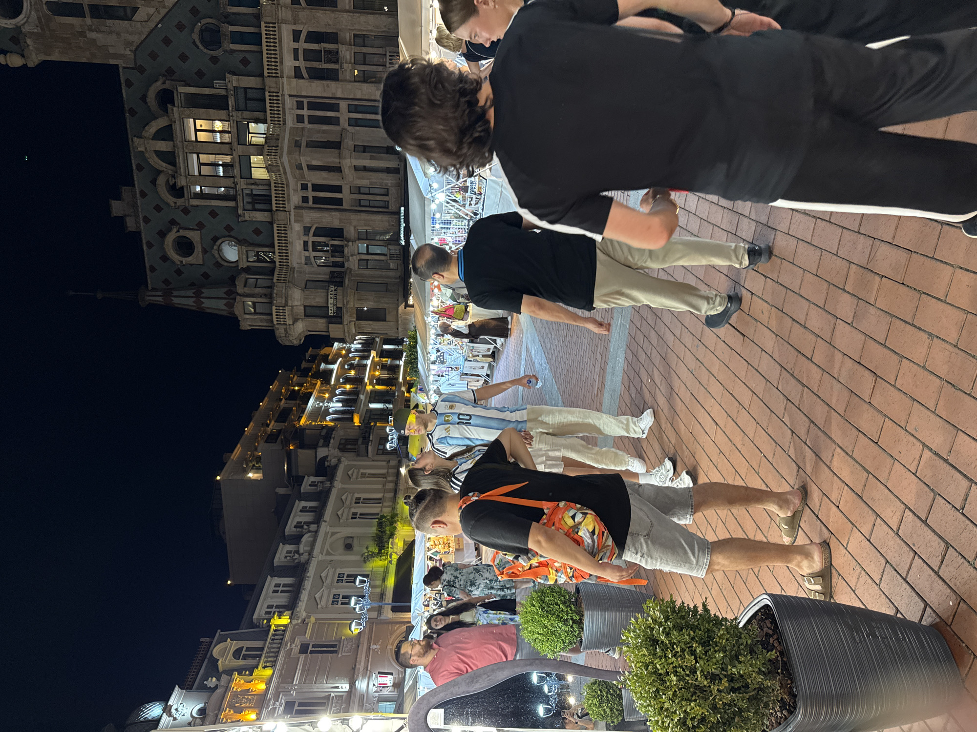 In Batumi, Georgia, at night, a group of people are walking along a brick-paved pedestrian street lined with buildings. The buildings have ornate architecture with illuminated windows and balconies. In the foreground, several individuals are visible. A man in a red shirt stands near a large potted plant and a mirror. To his right, a man wearing a black t-shirt and khaki pants walks away from the camera. Behind him, a man in a black t-shirt with white stripes on the sleeves is seen from the back. To the left, a man in a light-colored t-shirt and grey shorts, carrying an orange backpack, walks away from the camera. He is wearing brown sandals. To the right of the man in the red shirt, a woman in a white dress and a man in an Argentina soccer jersey are walking away. In the background, there are stalls or booths set up, possibly for a market or fair, with lights illuminating the area. The overall scene depicts a lively evening atmosphere in a city square.