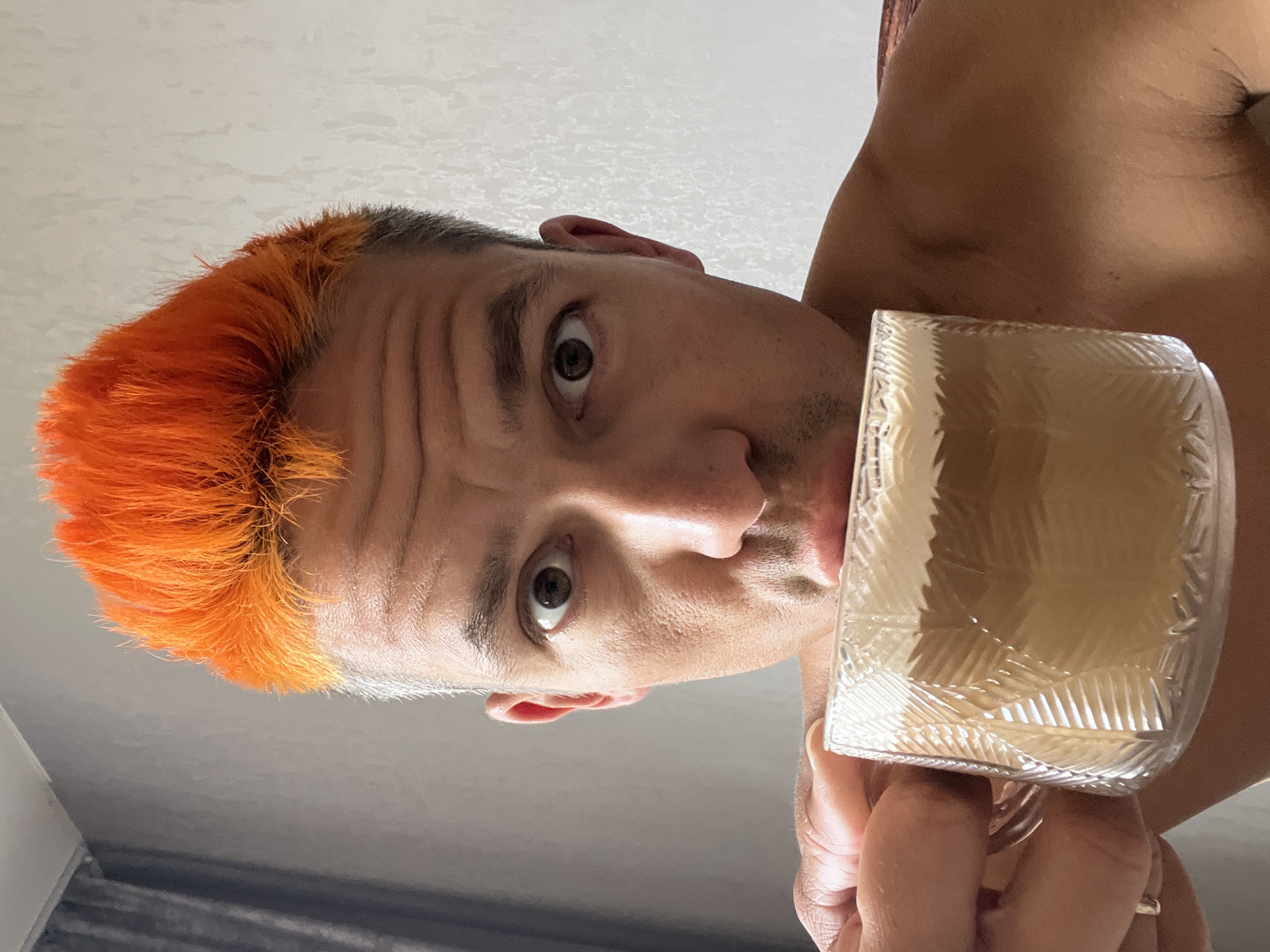 A man is holding a clear glass container filled with a light-brown liquid, positioned slightly below his face. He is looking directly at the camera. His hair is short, with the front dyed a bright orange. The background consists of a plain white wall or ceiling and a dark grey curtain. It is not possible to identify if the image was taken in the city of Chaikovsky, Russia.