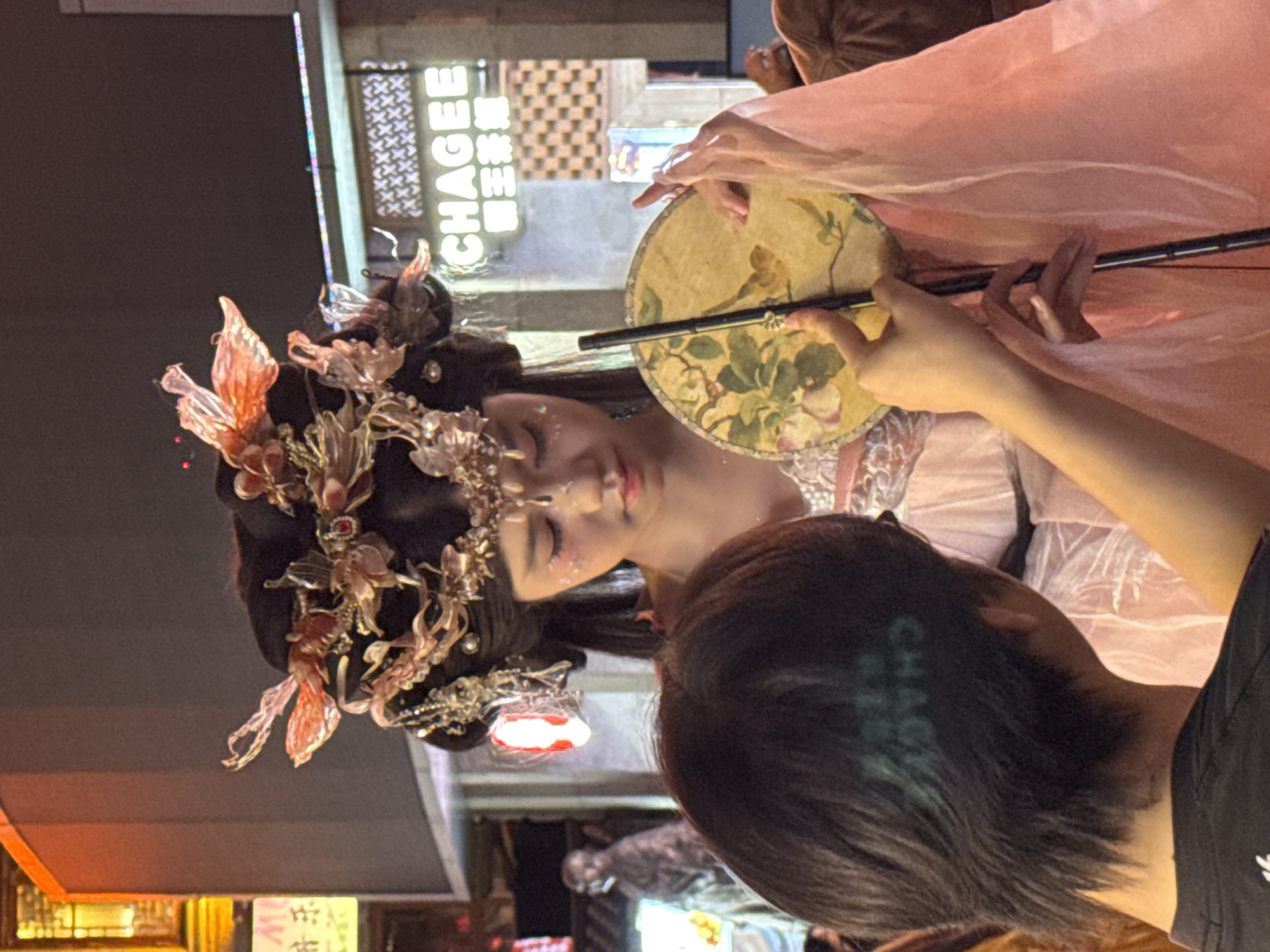 In Yuzhong District, China, a young woman adorned in traditional Chinese attire, complete with an elaborate headdress and makeup, poses for a photograph. She wears a light pink dress with sheer sleeves and holds a round fan decorated with a floral and bird motif. Her gaze is directed downwards, giving her a contemplative expression. A person, whose back is to the camera, is visible in the foreground, their dark hair partially obscuring their head. They appear to be holding the fan's handle, possibly assisting with the photoshoot. The background is an urban setting, featuring neon signs with Chinese characters and illuminated building facades, suggesting a lively street scene at night. A statue of a person in a contemplative pose is visible in the far left background.