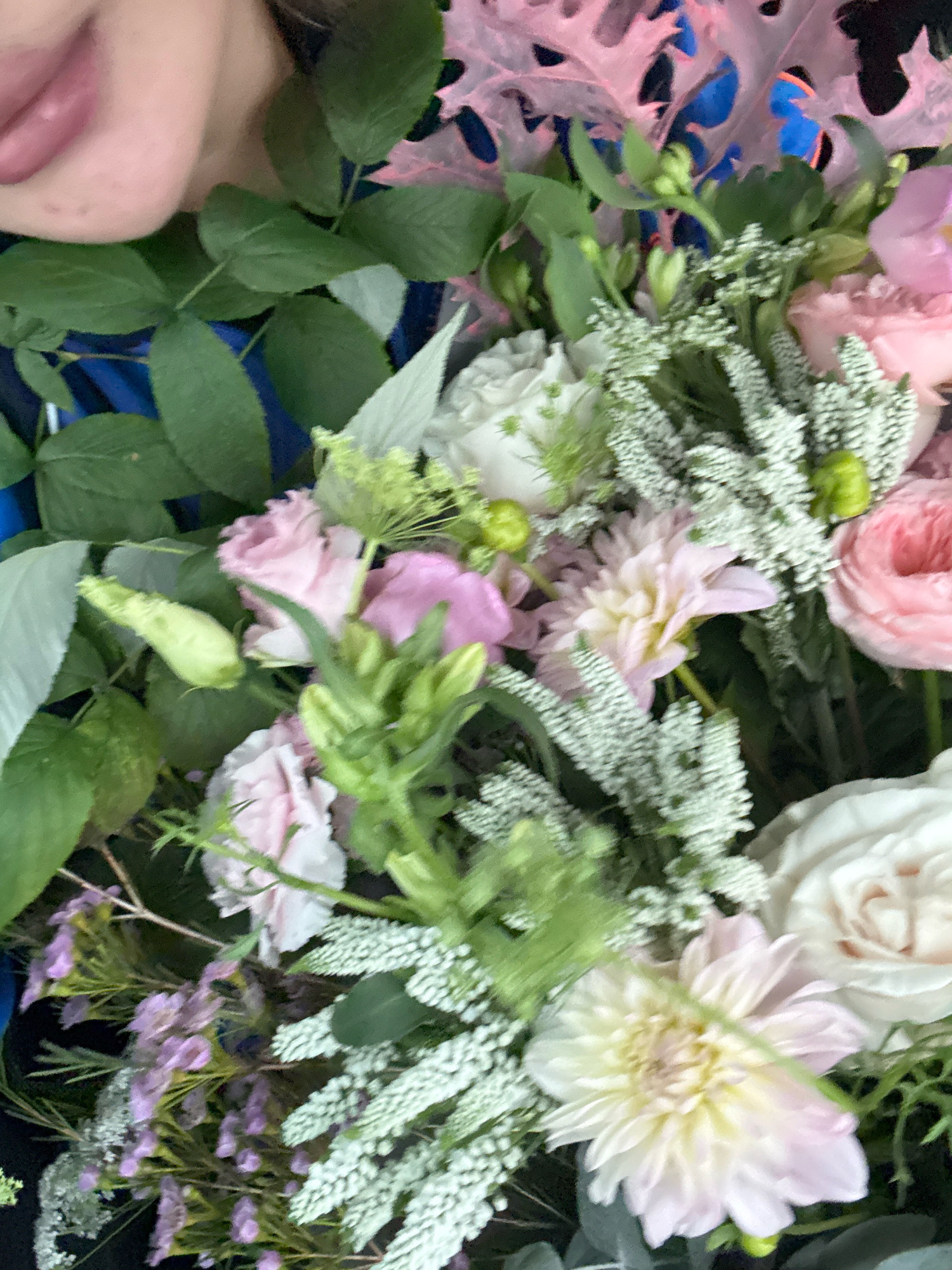 The image shows a close-up of a bouquet of flowers, primarily in shades of pink, white, and green. The bouquet contains various types of flowers and foliage, including roses, dahlias, and smaller, delicate white and purple blossoms. The flowers are arranged tightly together. In the upper left corner, a person's lips and chin are visible, with light-colored skin. The background is blurred, but appears to be dark with hints of blue and pink. There is no discernible location within the city of Tallinn, Estonia, or any specific environmental details that would suggest a particular place. The image focuses solely on the floral arrangement and a portion of a person's face.