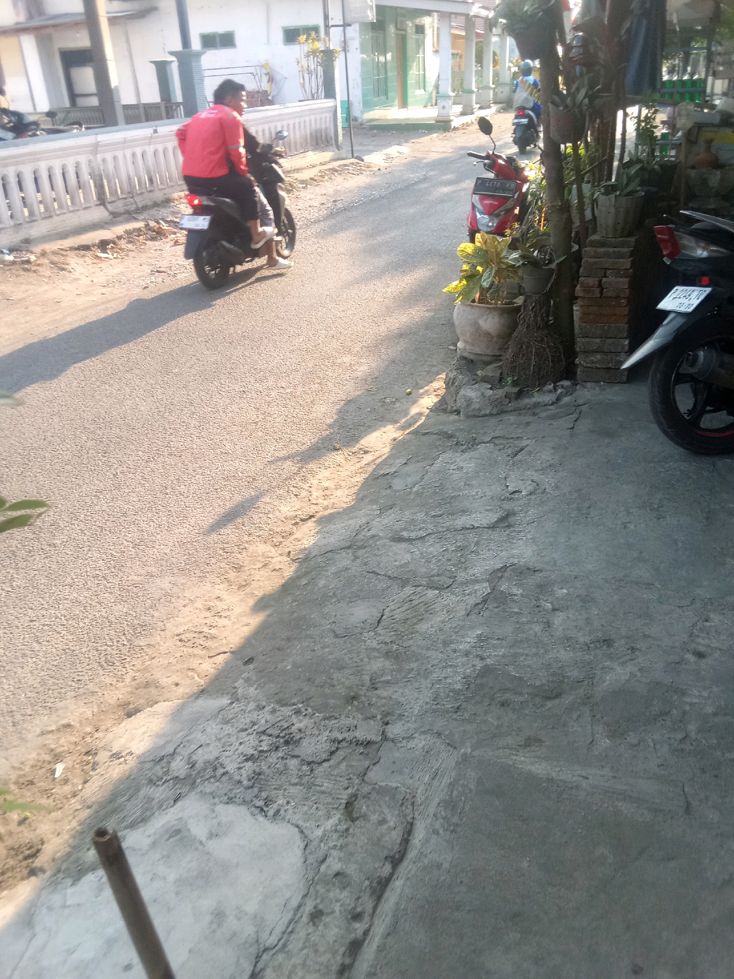 In the city of Jubung, Indonesia, a person wearing a red jacket rides a black motorcycle, with another person sitting behind them. The motorcycle is on a paved road that appears to be in a residential area, with houses and other motorcycles visible in the background. The sun is casting long shadows on the road, indicating it is either early morning or late afternoon. A concrete sidewalk runs along the right side of the road, featuring several potted plants and stacks of bricks. Another black motorcycle is parked on the sidewalk. The overall scene depicts a typical day in this Indonesian neighborhood.