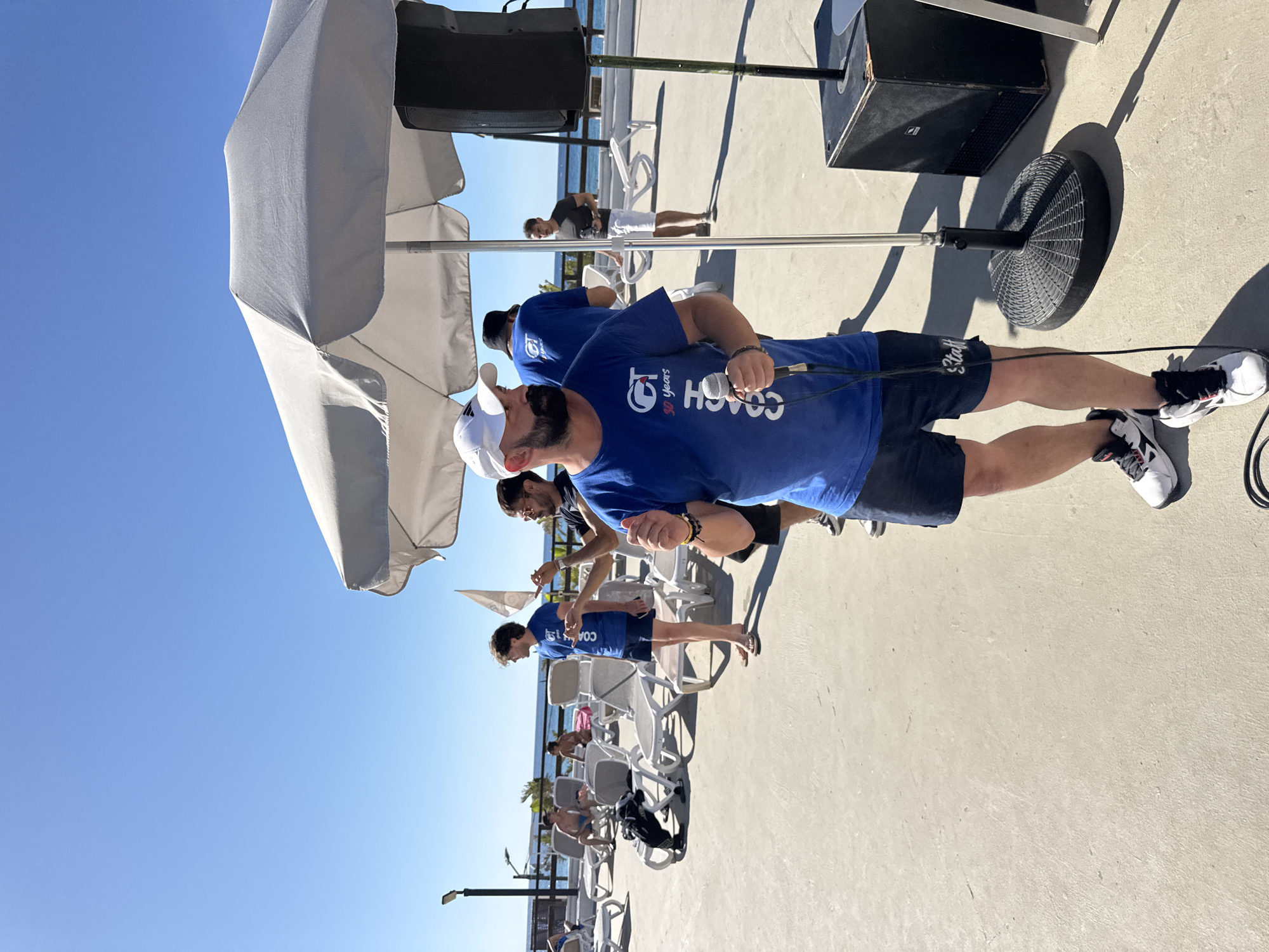 In Capaci, Italy, on a sunny day, a man wearing a blue t-shirt that says "CT COACH 30 Years" and a white baseball cap is holding a microphone and speaking. He is standing on a paved outdoor area with lounge chairs and umbrellas in the background. Behind him, other people are visible, some sitting on lounge chairs and others standing. The scene appears to be at a poolside or beachfront location, with the sea visible in the distance. A large black speaker is positioned next to a silver microphone stand and a large white umbrella. The overall atmosphere suggests an outdoor event or gathering.