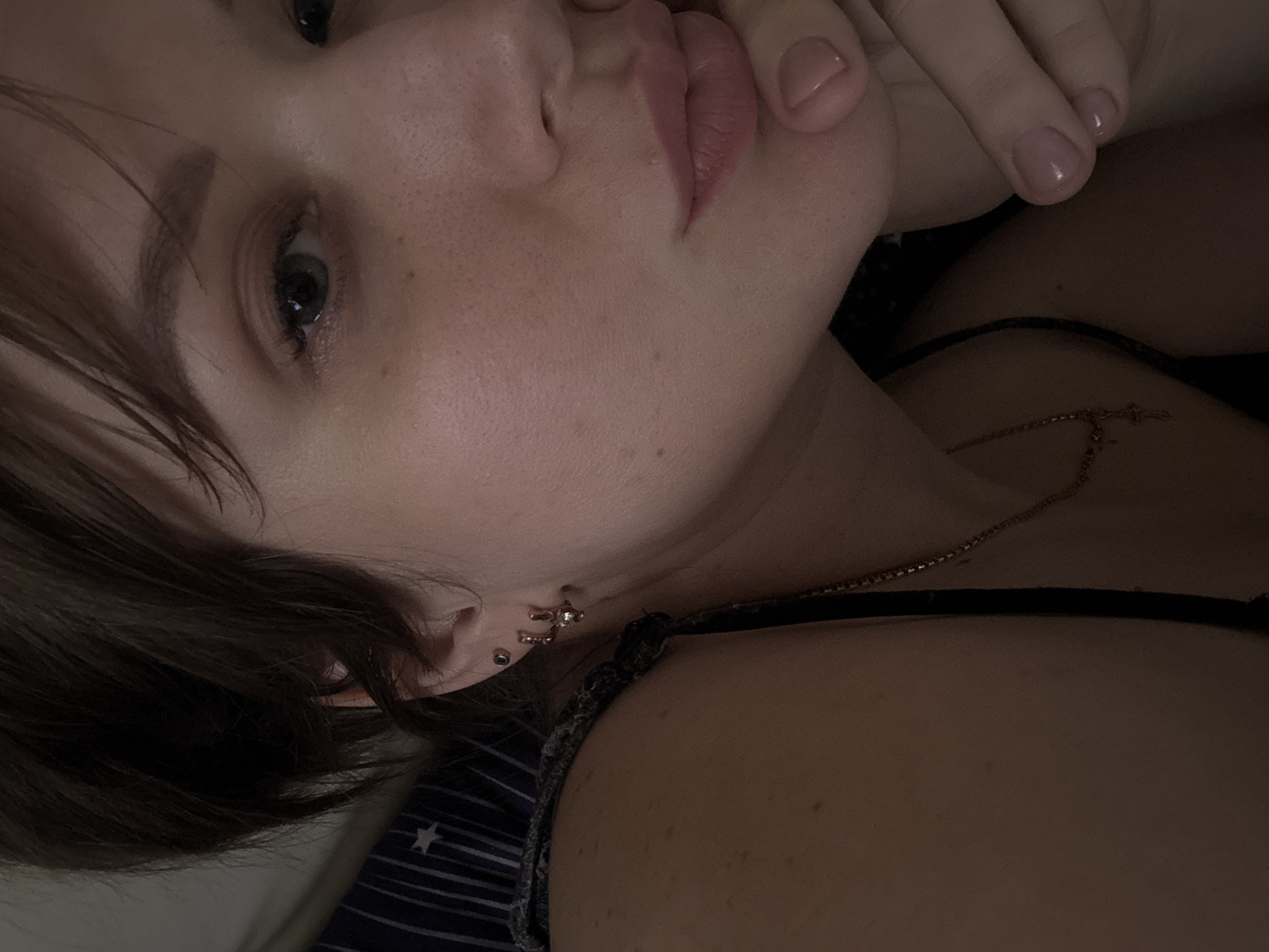 A close-up, dimly lit portrait of a young woman with fair skin, freckles, and short brown hair is angled slightly. Her left eye is visible, appearing grey-green, and her lips are pursed. Her right hand is partially visible, with neatly manicured fingernails, resting near her face. A delicate gold necklace is visible on her chest. She wears a black top with a narrow strap. Two ear piercings are visible on her left ear; one with a simple stud and another with a decorative earring. The background suggests she is lying on a bed with a patterned blue and white bedsheet visible. There is no information to suggest the location is Chaikovsky, Russia.