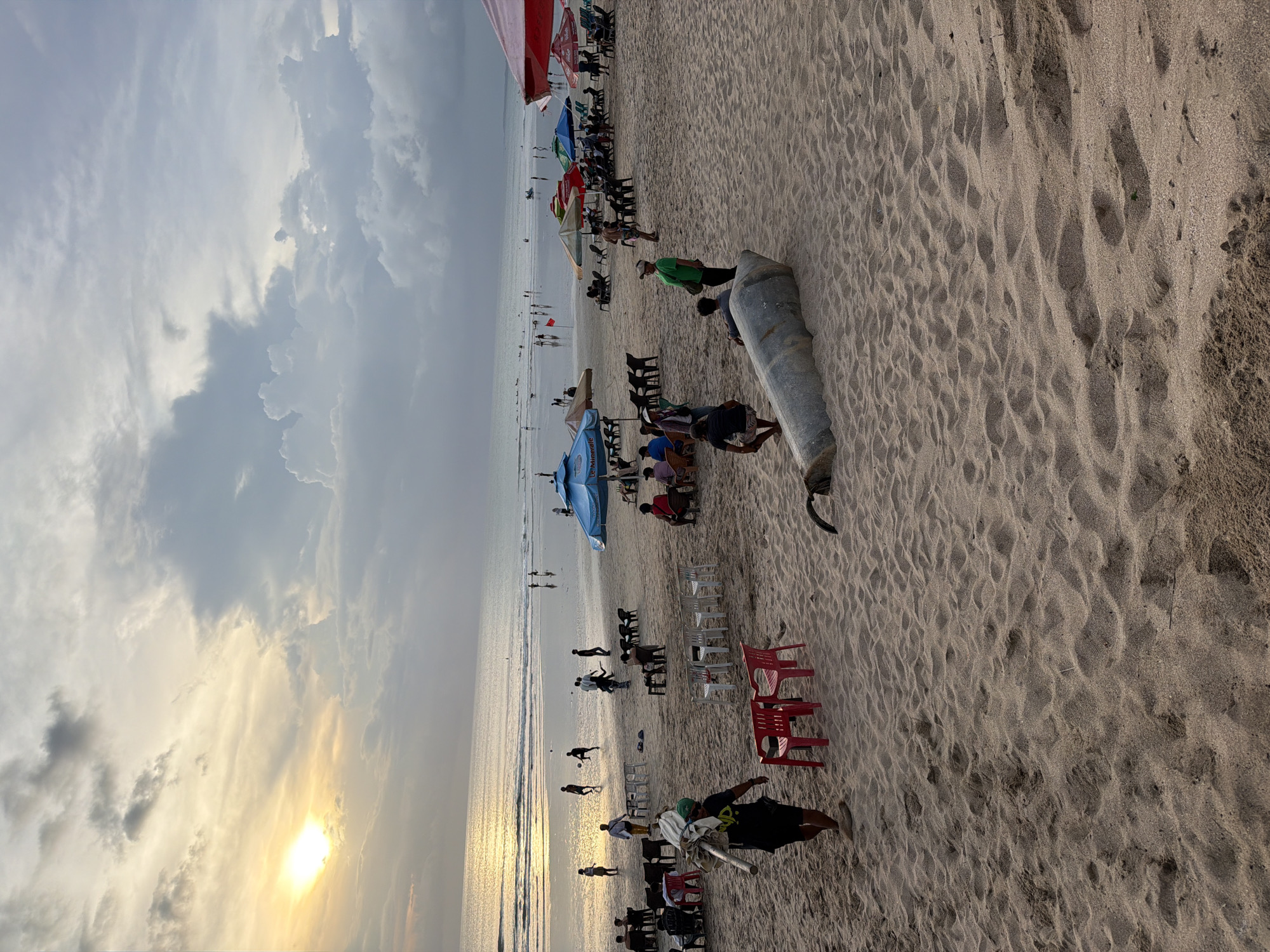 The image shows a crowded beach at sunset in Kuta, Indonesia. The sky is filled with clouds, and the sun is low on the horizon, casting a golden glow over the scene. Many people are on the beach, some sitting in chairs under umbrellas, others walking along the shoreline or swimming in the ocean. There are numerous beach chairs, some in rows, and a few red plastic chairs near the foreground. A metal object is on the sand, with people standing nearby. The sand is textured with footprints and patterns.