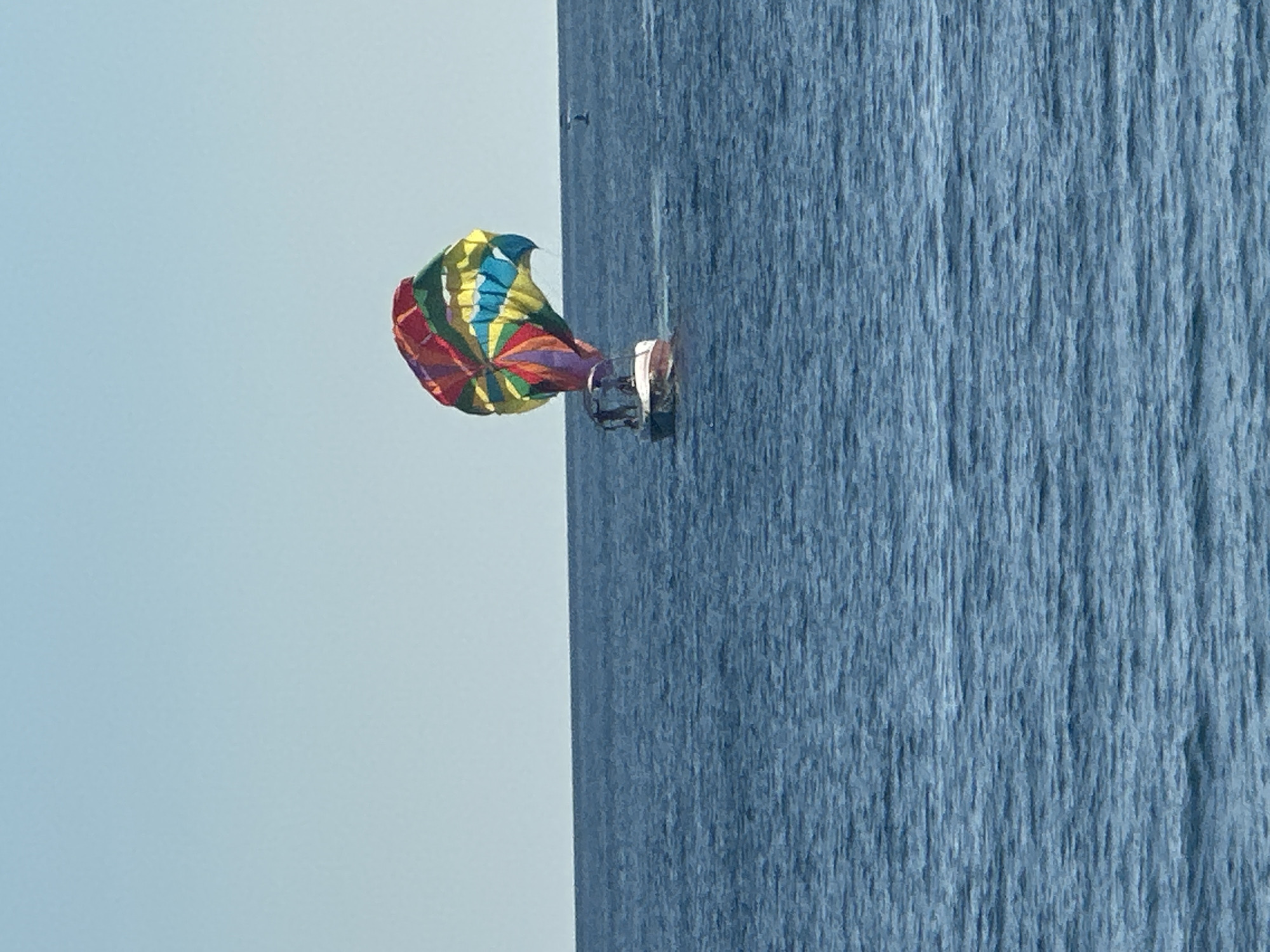 The image depicts a boat with a deployed parasail on the open water near Budva, Montenegro. The sky, occupying the upper portion of the frame, is clear and light blue. The lower two-thirds of the image show calm, dark blue water with discernible ripple patterns. A white motorboat, featuring red accents and a stern platform, is positioned slightly right of center, moving generally away from the viewer and towards the right side of the frame. Two indistinct human figures are visible standing on the boat's stern. Directly behind the boat, a large, inflated parasail is airborne, exhibiting prominent segments of red, yellow, green, and blue fabric. A small, dark, unidentifiable object is faintly visible on the distant water surface near the right horizon.