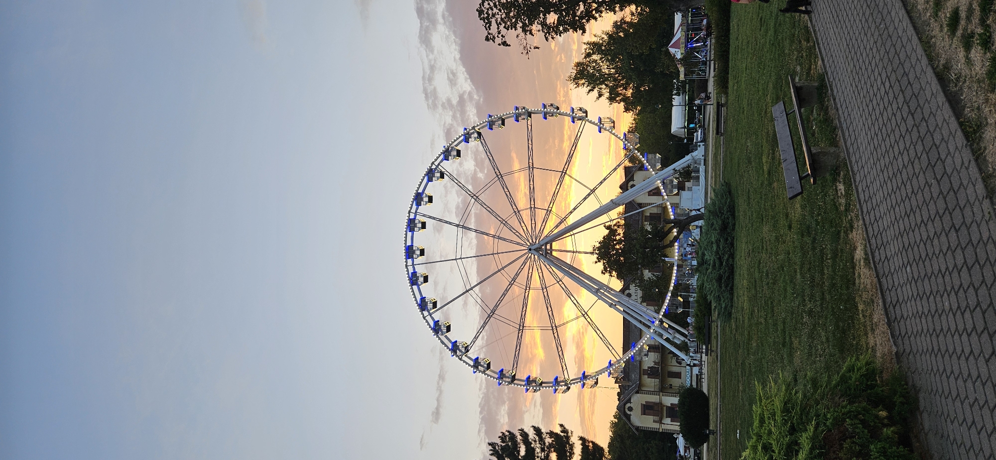 The image captures a sunset view of a Ferris wheel in the city of Keszthely, Hungary. The Ferris wheel, illuminated with blue and white lights, stands tall against a sky painted with vibrant orange and pink hues from the setting sun. In the background, a building with a sign that reads "BALATON" is visible, hinting at its proximity to Lake Balaton. The foreground features a grassy park with trees and bushes, a paved walkway, and a wooden bench. Several people are present in the scene, some walking or standing near the Ferris wheel and other amusement park attractions, suggesting a lively evening atmosphere. The overall scene depicts a recreational area at dusk, with the Ferris wheel as the prominent centerpiece.