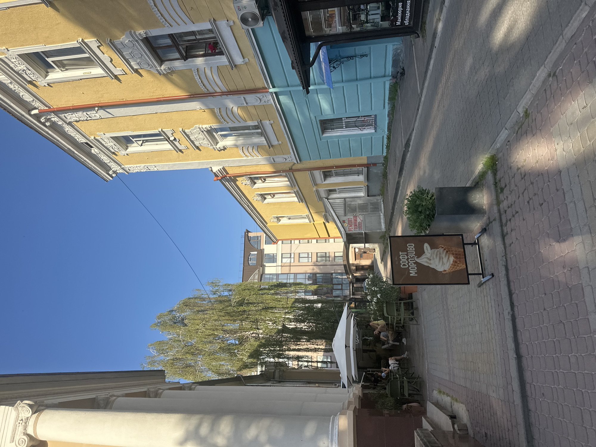 The image depicts a narrow street scene in Ternopil, Ukraine, bathed in bright sunlight. On the left, a building with large white columns lines the sidewalk, leading to a cafe with white umbrellas and patrons seated at tables. To the right, a vibrant yellow building with intricate white stucco detailing stands prominently, its lower half painted a striking turquoise. A street sign advertising "Soft Ice Cream" with an image of a cone is placed on the cobblestone sidewalk. Further down the street, other buildings are visible, including a modern multi-story structure. The sky is clear and blue.