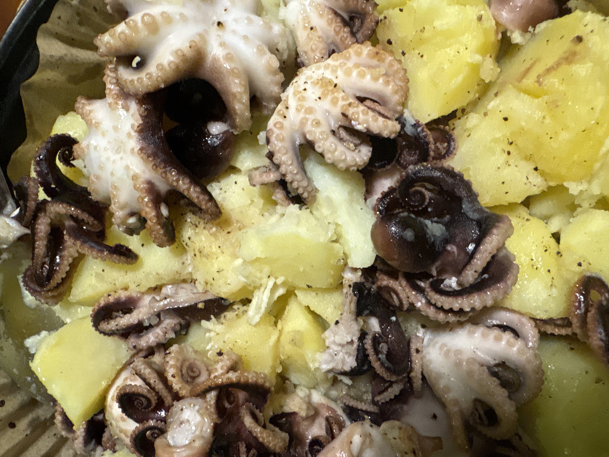 The image shows a close-up, overhead view of a dish containing cooked baby octopuses and chunks of yellow potato. The octopuses are small, with visible tentacles and suction cups. Some are a light pinkish-white color, while others are a darker purplish-brown. The potatoes are cut into irregular pieces and appear to be seasoned with black pepper. The dish is contained within a dark-colored, possibly metal or ceramic, baking dish lined with what appears to be parchment paper. The overall impression is of a rustic, home-cooked meal. There is no information in the image to indicate a specific location in Nha Trang Ward, Vietnam.