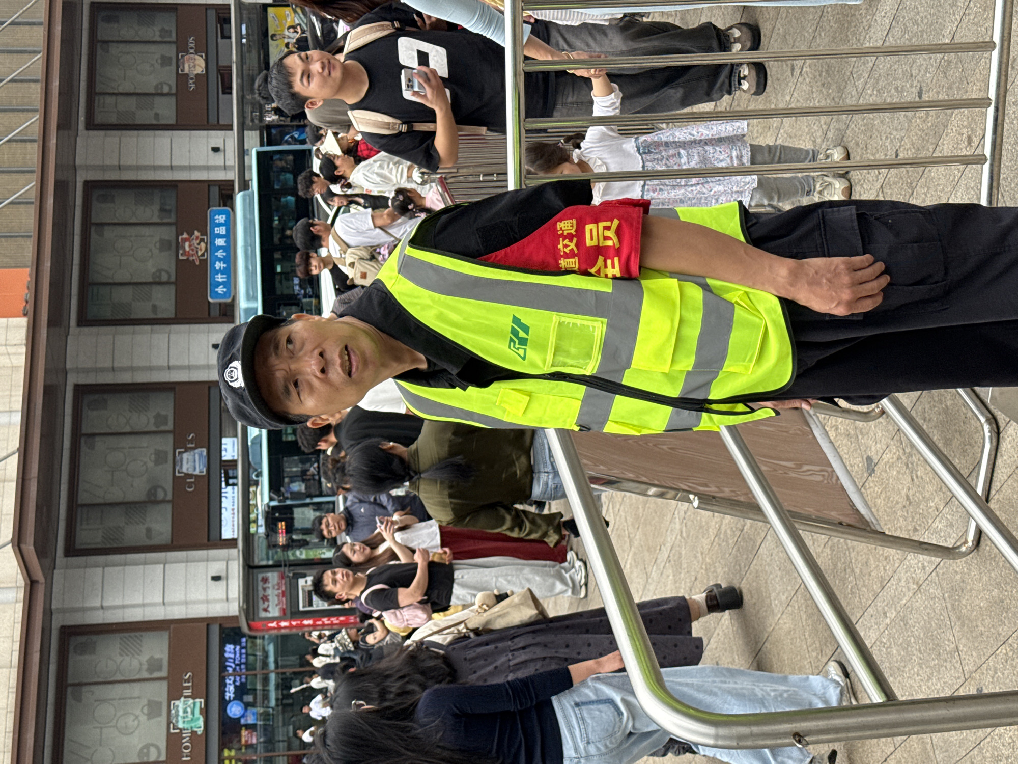 The image captures an outdoor scene in Guang'an, China, featuring a man wearing a yellow safety vest and a black cap with a badge. He has a red armband with Chinese characters, standing behind a metal railing. Behind him, a crowd of people stand and sit. In the background, there is a bus, and storefronts are visible, one labeled "HOME TILES" and another reading "SPORTS GOODS" in English. Signage includes one for "小什字小商品站". The image is taken at a close range, focusing on the man in the safety vest.
