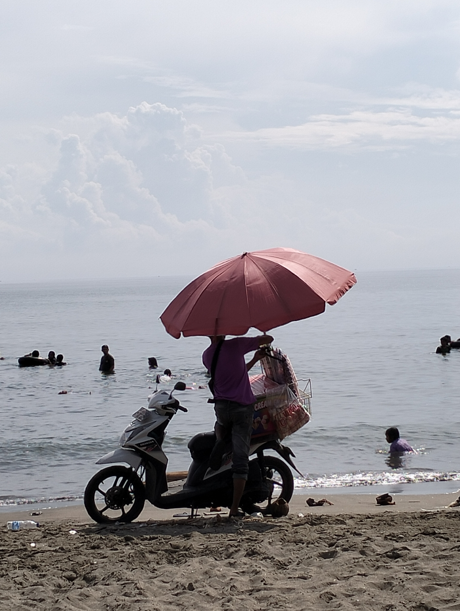 On a sandy beach in Lhokseumawe, Indonesia, a person wearing a purple shirt and gray jeans stands behind a white Honda scooter, holding a large, pink umbrella. A wire basket filled with packaged goods is attached to the back of the scooter. Several people are in the water, some swimming and some standing. The sea meets the cloudy sky in the background. Debris litters the beach.