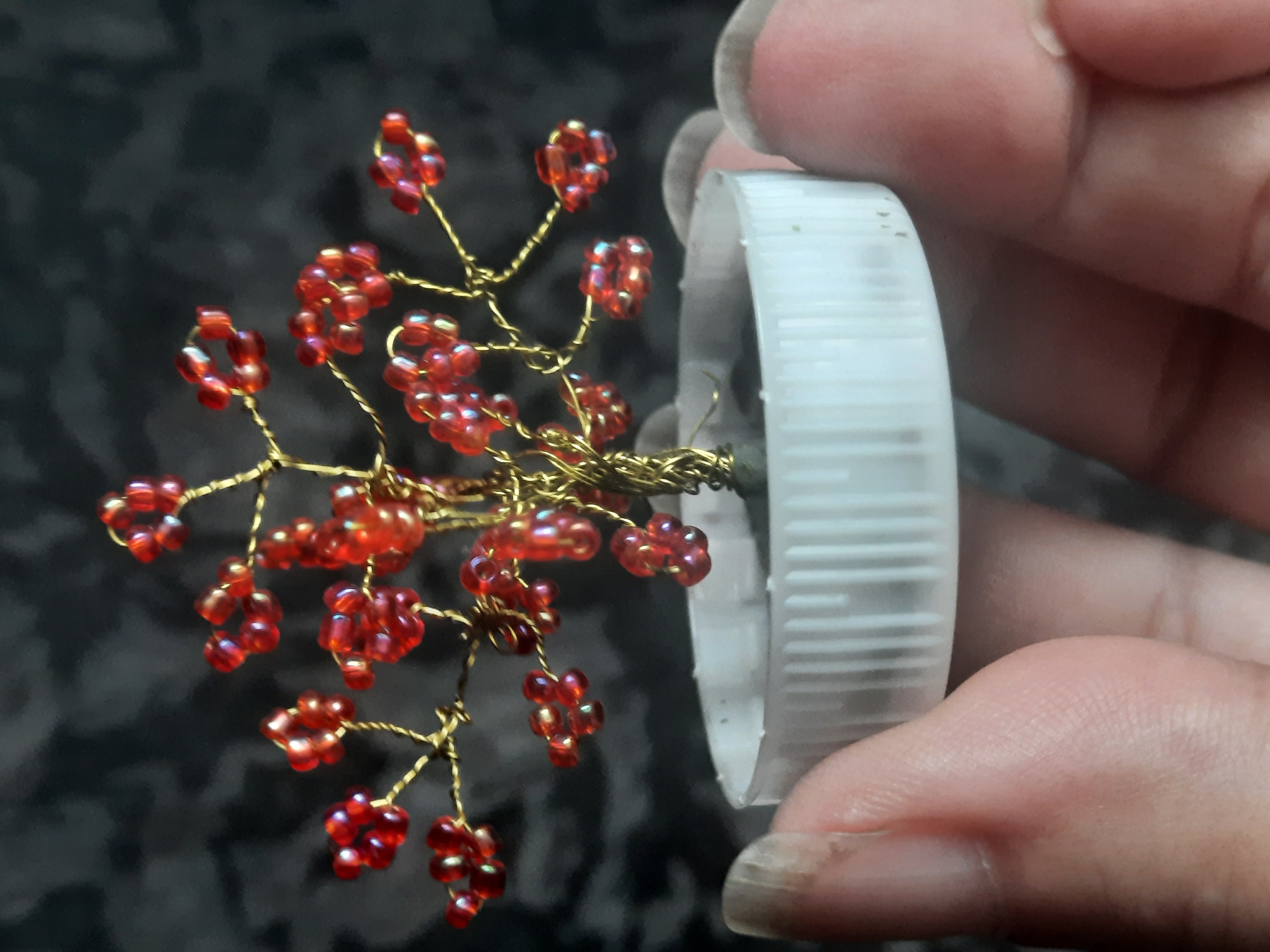 A close-up, eye-level shot focuses on a miniature beaded tree crafted with red beads and gold wire. The tree is rooted in what appears to be a white bottle cap, which is held between two hands. The background is a dark, textured surface, possibly fabric. The scene is likely indoors. The provided image does not contain any information about the location in the city of Sluck, Belarus.