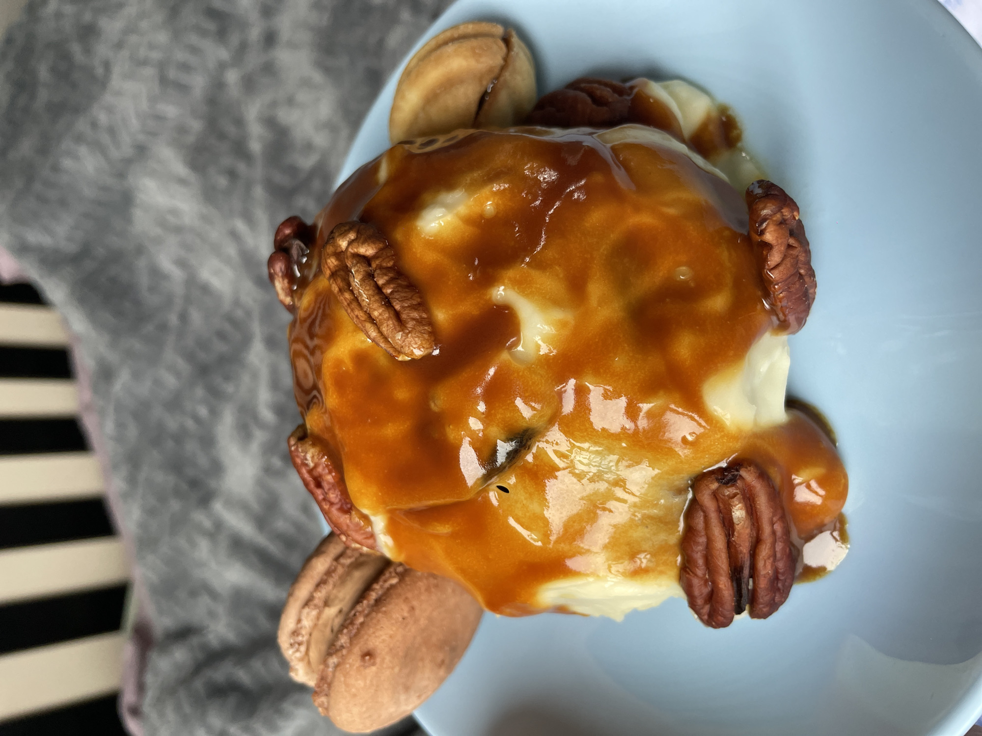 This image captures a close-up view of a dessert, positioned on a light blue ceramic plate, indoors in Sochi, Russia. The primary subject is a rounded food item generously coated in a viscous, amber-hued caramel sauce, which exhibits a reflective surface. Multiple whole pecan halves are affixed to the surface of the caramel and distributed around the base of the central item. A light brown macaron with a textured surface is visible on the left side of the plate, partially obscured. On the right side, a small, light brown, bivalve-shaped cookie is present. The light blue plate rests upon a crumpled dark gray fabric. The background behind the fabric is out of focus, revealing vertical alternating dark and light stripes, suggesting a piece of furniture or wall decor. No people are visible, and no actions or interactions are depicted.