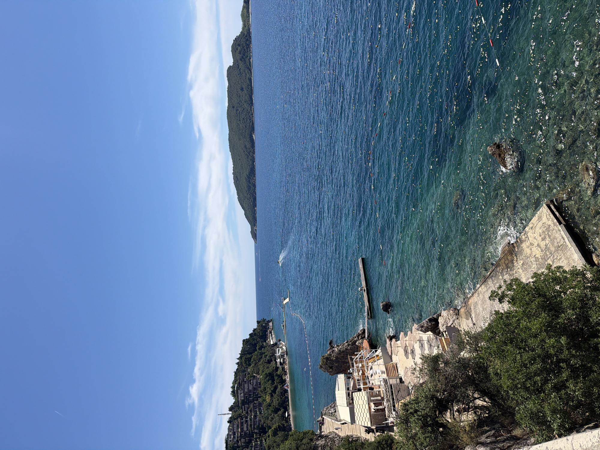 This is a high-angle, full-shot, vertical image of a coastal scene in Budva, Montenegro. The foreground features a concrete walkway and a cluster of umbrellas on a rocky area adjacent to the sea. A building with a white canopy and open sides, possibly a cafe or restaurant, is situated on the left, with a few visible patrons. Lush green trees and bushes grow on the rocky embankment.
The sea dominates the middle and right portions of the image, displaying a gradient of blue hues from turquoise near the shore to a deep indigo further out. Numerous small buoys, some red and white, are scattered across the water, marking a swimming area. A wooden pier extends into the sea from the rocky shoreline, with a person visible on it. In the distance, a small boat leaves a wake on the water, and a larger island covered in dense green vegetation sits on the horizon.
The background shows a hillside densely populated with modern, terraced buildings, also covered in greenery, overlooking the bay. The sky is a clear, bright blue with a few wispy white clouds and a faint contrail from an aircraft. The overall impression is a sunny day at a scenic coastal resort.
