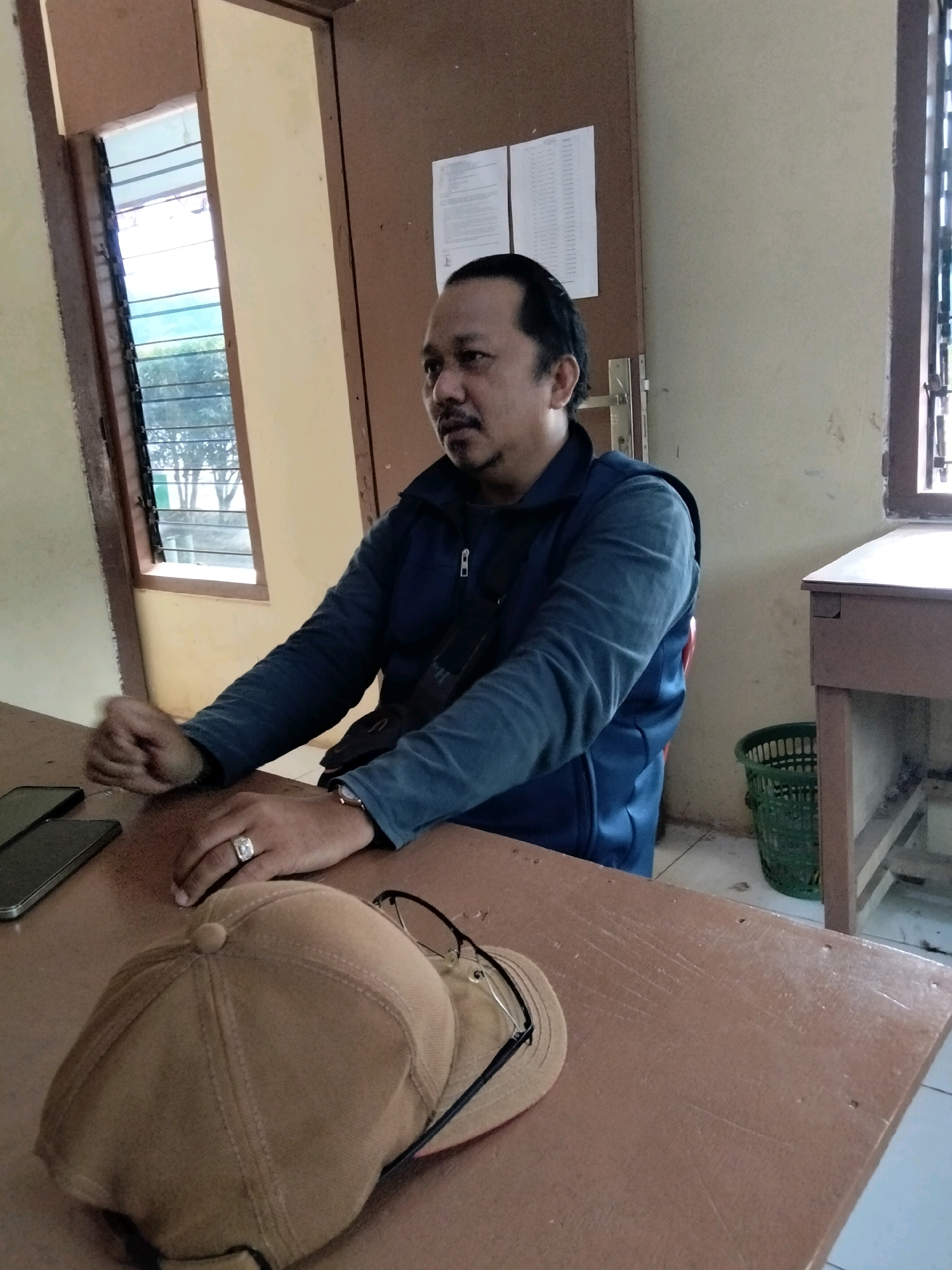 A man with dark hair and a mustache is seated at a wooden table, facing slightly to the right. He is wearing a dark blue jacket over a lighter blue shirt and has a ring on his left ring finger. Two smartphones are on the table in front of him, along with a tan baseball cap and a pair of eyeglasses. The room has beige walls and a wooden door with papers pinned to it. A window with horizontal blinds is visible on the left, showing trees outside. A small green trash can is situated near a wooden desk on the right side of the room. The scene appears to be an indoor office or classroom setting. There is no specific indication of the city of Cinta Manis Lama, Indonesia.