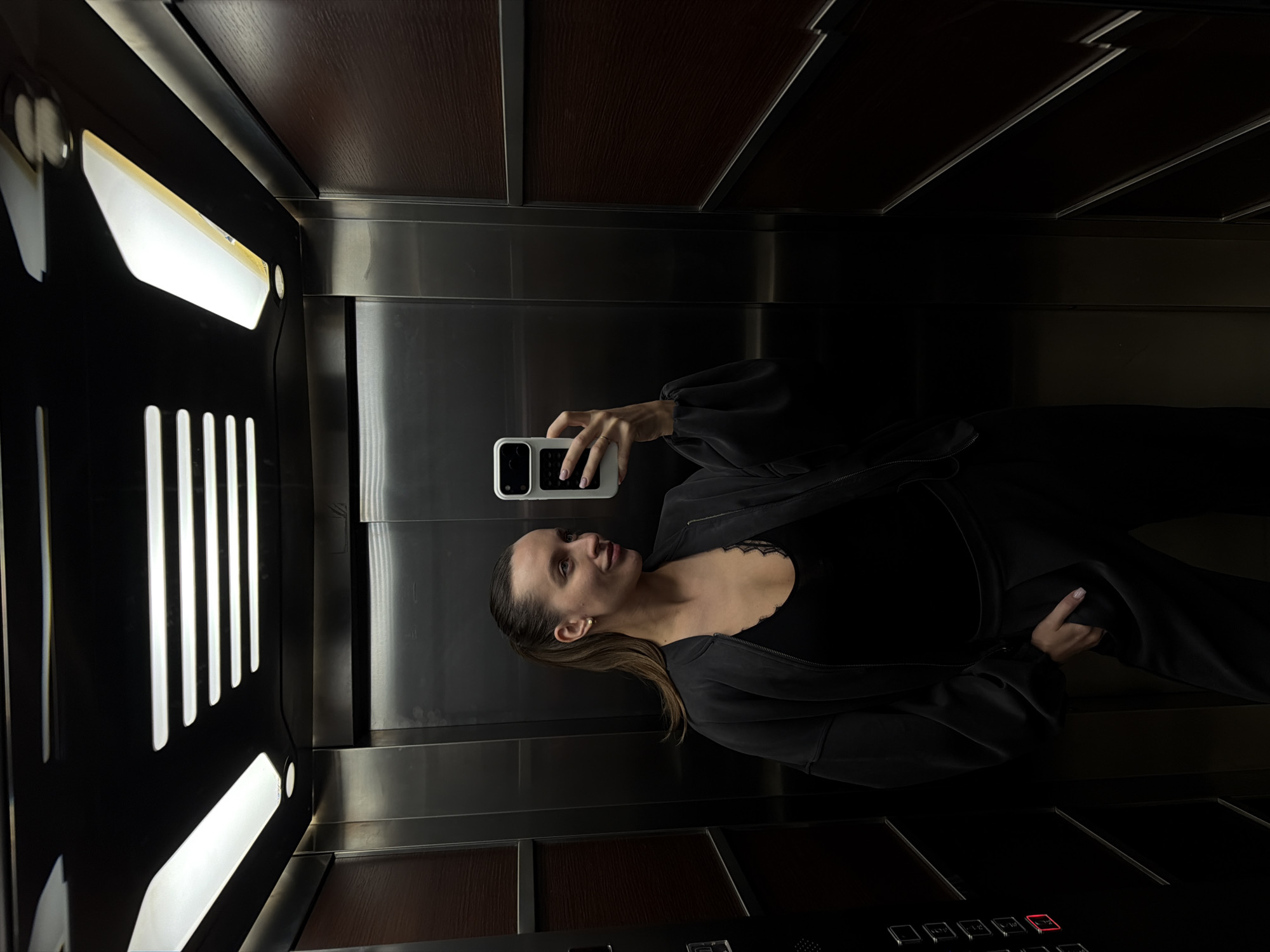 A woman is taking a selfie in an elevator. She is wearing a black top with lace detailing and a black jacket. Her hair is slicked back and she is holding a white smartphone in her right hand. The elevator has stainless steel walls and a dark wood panel on top. The elevator's control panel is visible at the bottom, and a light fixture is on the left. The image was probably taken in Budva, Montenegro, but there are no specific landmarks visible to confirm this.
