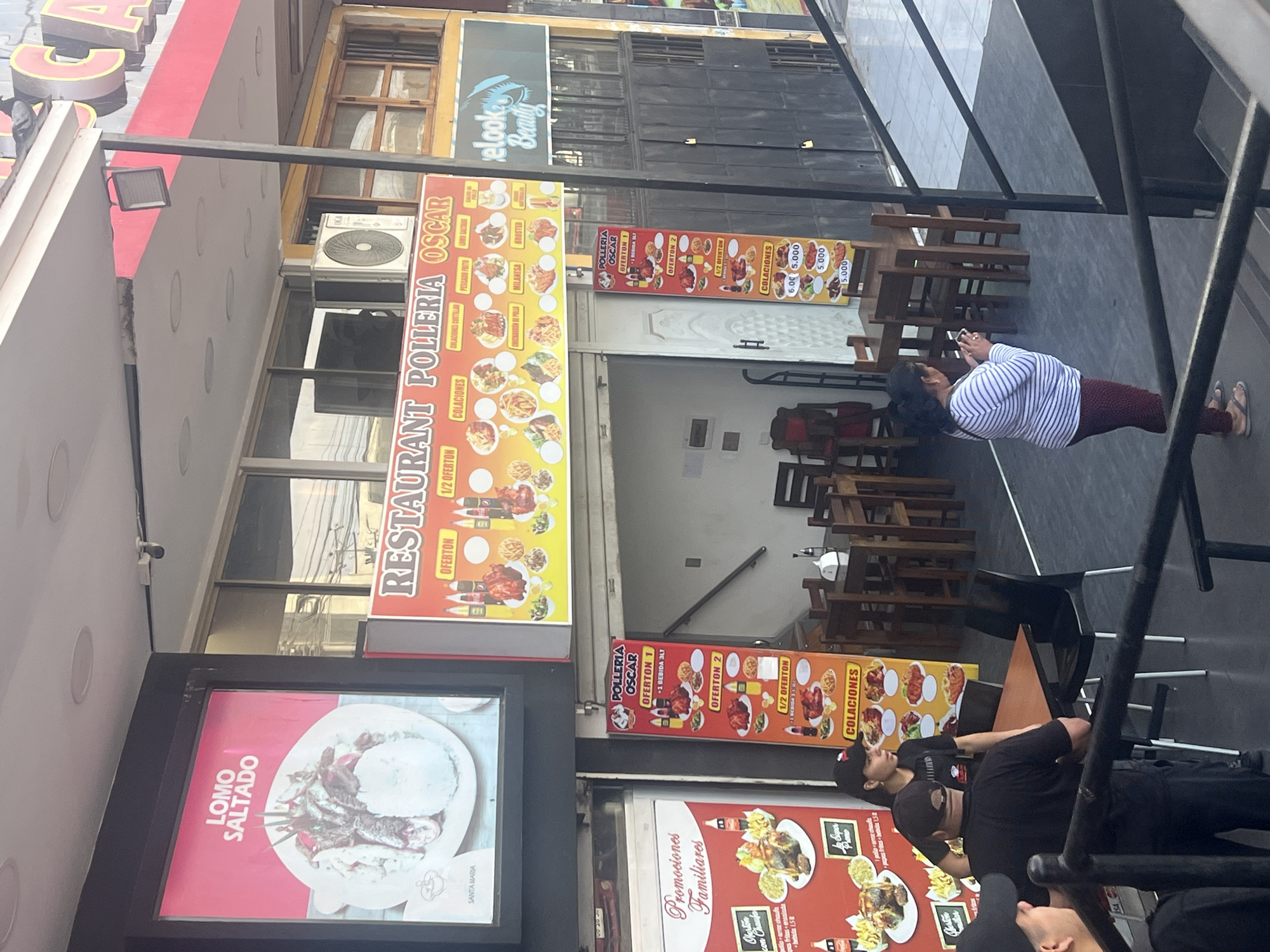 The image shows the exterior of "RESTAURANT POLLERIA OSCAR," a restaurant in what appears to be Arica, Chile, according to the OCR in the image. The restaurant has a bright yellow and red sign with menu items displayed. Banners advertise special offers, "OFERTON 1" and "OFERTON 2." A large sign for "LOMO SALTADO" is also visible. Several people are present. One woman wearing a striped shirt and maroon pants appears to be taking a photo, while three individuals stand nearby and are wearing black and dark clothing, suggesting they may be staff. The environment includes outdoor seating and parts of surrounding buildings, including a beauty salon called "elook Beauty."