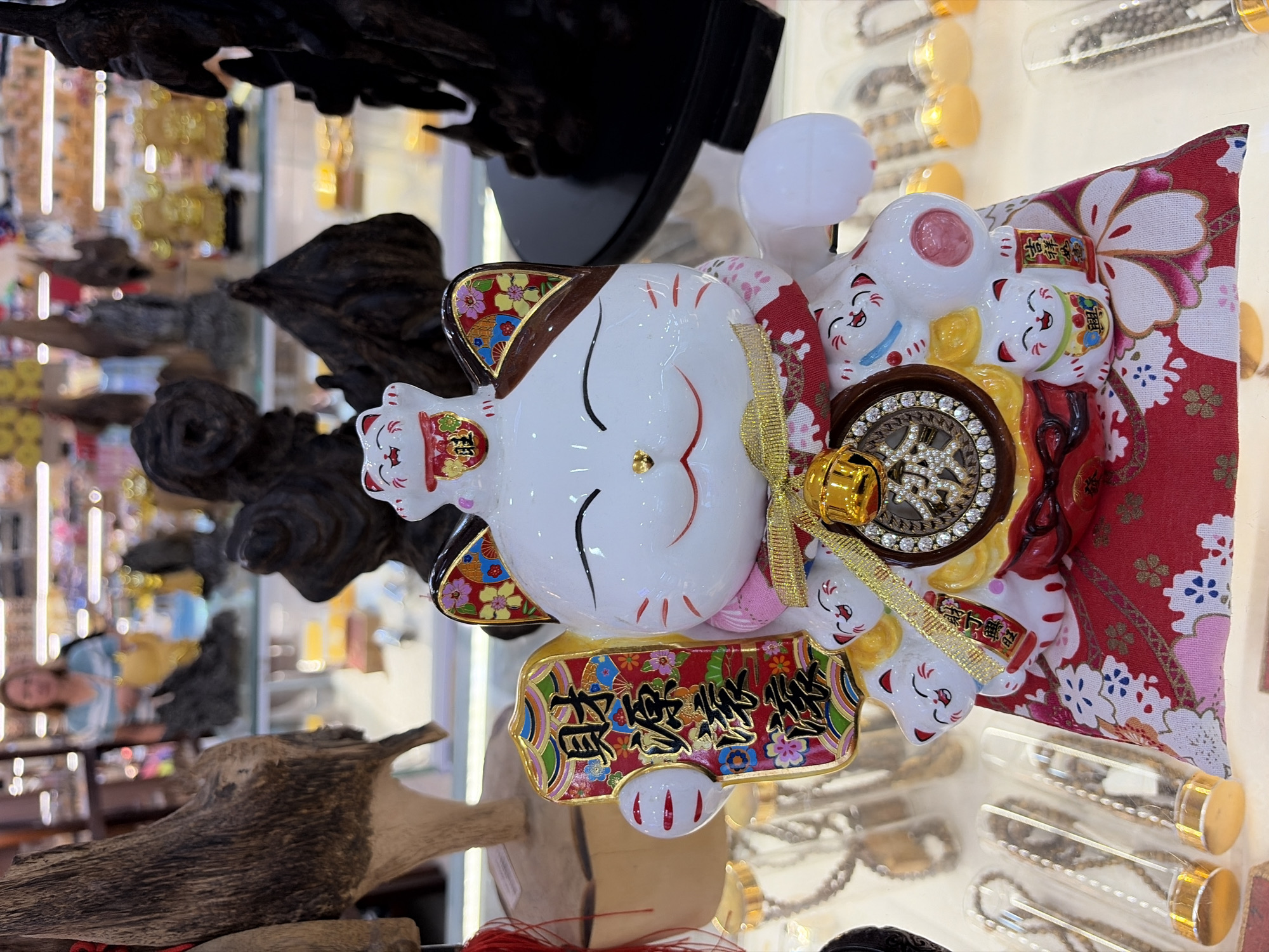 A Maneki-neko, or "beckoning cat" figurine, is prominently displayed in what appears to be a shop in Da Nang, Vietnam. The figurine is white with brown markings on its ears and eyes, and it is adorned with red and gold accents. It holds a decorative plaque with Chinese characters and is decorated with smaller cat figurines and floral patterns. The figurine sits on a red cloth with a floral design. In the background, shelves are filled with various goods, including what look like wooden carvings and other decorative items. A woman is visible in the background, out of focus. The lighting suggests an indoor environment, likely a retail setting.