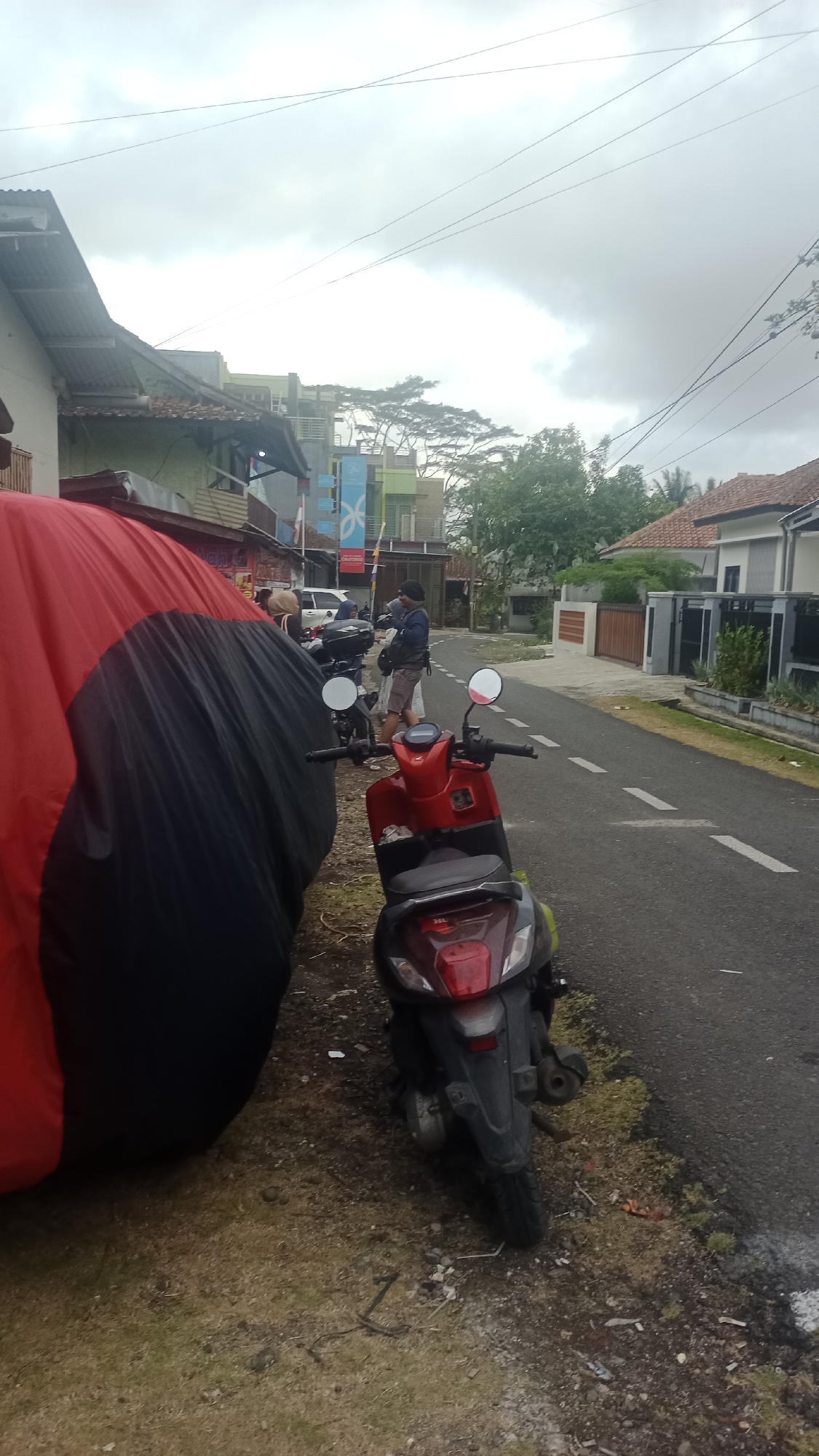 In Pakemitan, Indonesia, a street scene unfolds under an overcast sky. A large object covered by a red and black tarp dominates the left side of the frame, suggesting it is parked. To its right, two motorbikes are visible. One is a dark-colored scooter, partially obscured by the tarp. The other, a bright red scooter, is positioned closer to the road and faces away from the viewer, its rear lights clearly visible.
A person dressed in a dark jacket and shorts stands beside the motorbikes, seemingly engaged with them or preparing to depart. They are holding a phone. Further down the street, another individual, wearing a headscarf and a light-colored jacket, is also near the motorbikes, partially obscured. The street itself is paved, with dashed white lines marking the lanes. Houses with pitched roofs line the right side of the road, and various trees and foliage are interspersed throughout the environment. The overall impression is of a quiet, everyday moment in a residential area.