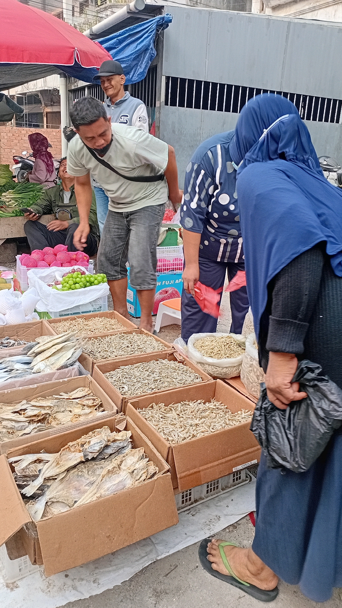 The image depicts an outdoor market scene in Jambi, Indonesia, likely a street-side stall. In the foreground, several cardboard boxes are filled with dried fish and small dried anchovies, typical produce for sale at such a market. A person wearing a navy blue headscarf and dress, along with black flip-flops, is holding a black plastic bag. To their left, another individual in a light-colored t-shirt and dark shorts is bending over, seemingly examining the goods. Behind them, a young man in a light blue and white jacket and a black cap is looking towards the camera with a slight smile. Further back, a person in a green jacket is seated, holding a mobile phone. The background shows a building with barred windows and a blue tarp overhead, suggesting a semi-permanent market setup. The ground appears to be paved or concrete, common in urban areas. The atmosphere suggests a typical day of commerce at a local Indonesian market.