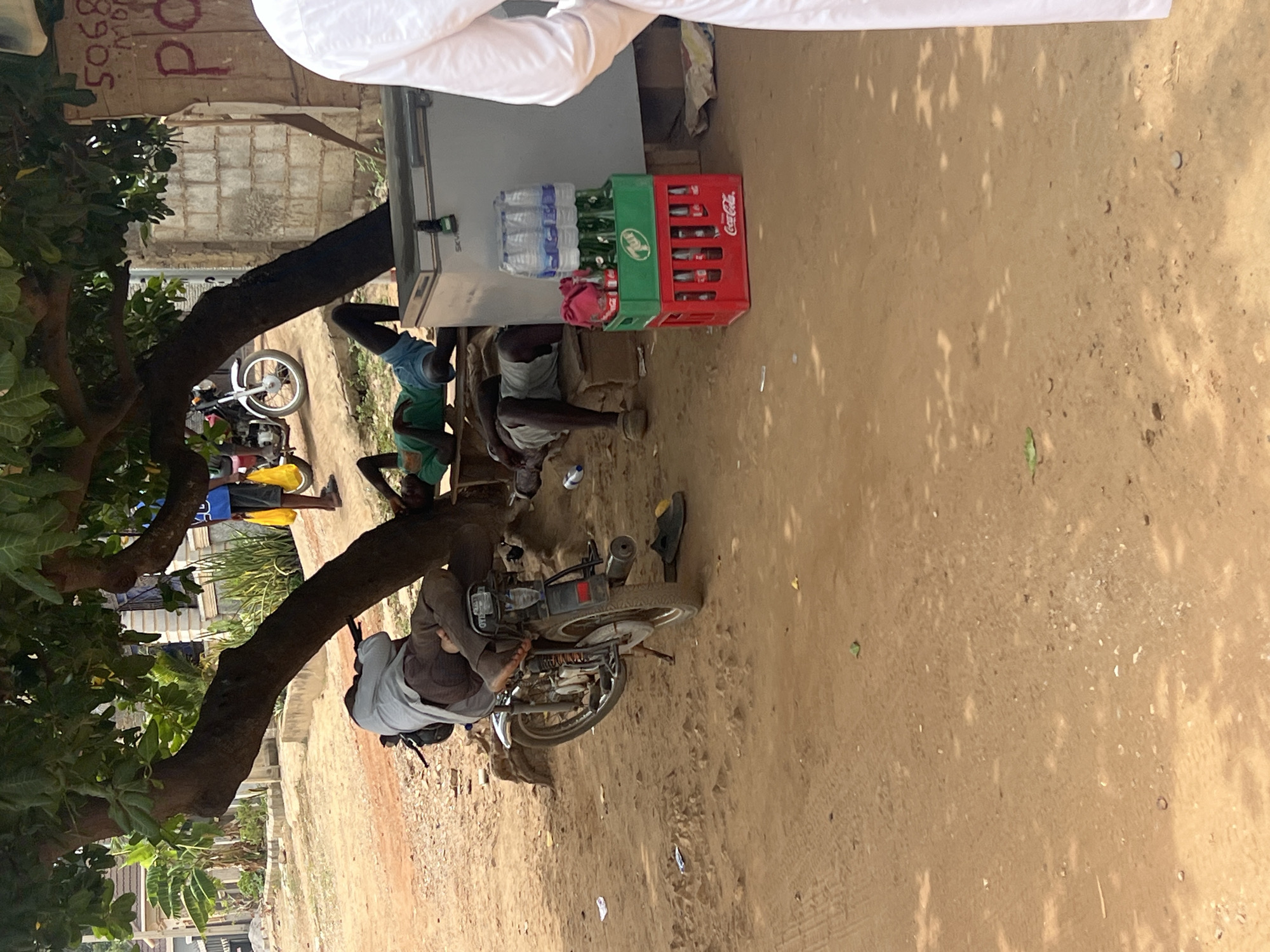 The image depicts a street scene in Abuja, Nigeria. A group of people are gathered around a refrigerator used as a makeshift shop, where bottled water, soda, and juice are stacked. A red Coca-Cola crate is visible on top. Some individuals are seated or leaning near the refrigerator, while others appear to be interacting with a parked motorcycle. The ground is sandy, and a tree provides shade. A building is visible in the background.