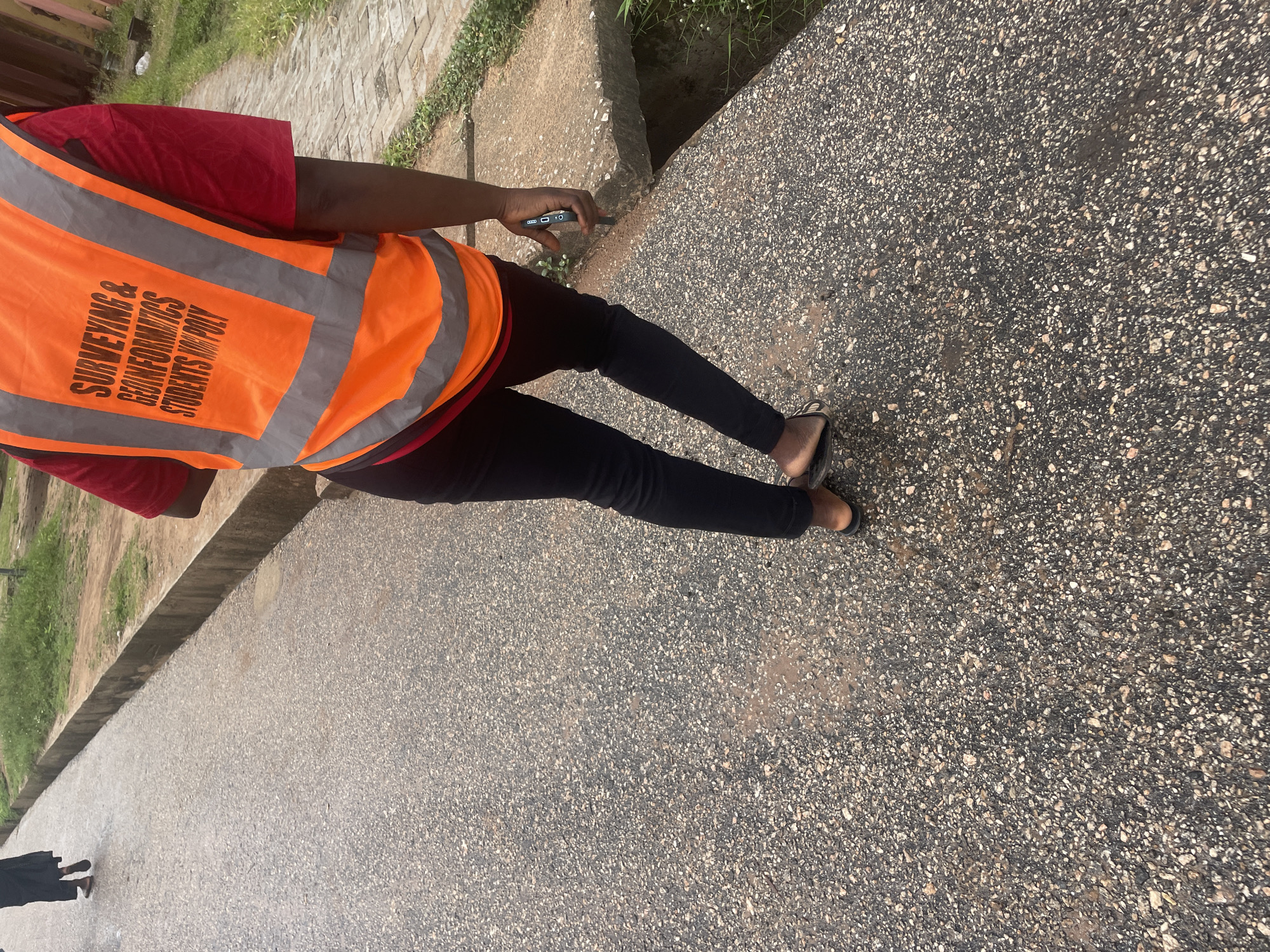 A medium shot captures the back of a person wearing a bright orange safety vest over a red shirt and black pants, as they walk on a paved asphalt road in Lokoja, Nigeria. The vest features reflective silver stripes and text that reads "SURVEYING & GEoinformatics STUDENTS KOGI POLY". The person is holding a black mobile phone in their right hand. To the left, a second individual, partially visible, walks away from the camera, wearing a black garment. The road has a rough, speckled texture, with scattered patches of lighter colored stones. A concrete drainage channel runs alongside the road on the right, with a section of it broken or collapsed, revealing a dark opening. The surrounding environment includes a grassy verge and some vegetation. The lighting suggests an overcast day, possibly after rain, as there are some wet patches on the asphalt.