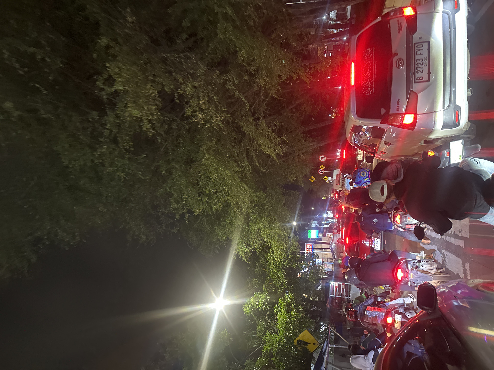 The image captures a congested traffic scene at night in North Jakarta, Indonesia. A dense line of motorcycles and cars is moving slowly or stopped on a brightly lit road. Pedestrians walk along the side of the road. A white car with the license plate "B 2723 FYO" is visible in the upper right corner, its brake lights illuminated. Additional license plates visible in the image are "B 4626 UEC" and "B 2054 K." The car has an inscription on the back that reads, "في الاء ربكما تكذبان" in Arabic. The image is taken from an elevated position, likely from inside a car. Trees and foliage line the left side of the road, with street lights providing illumination. Several traffic signs are also visible near the cars.