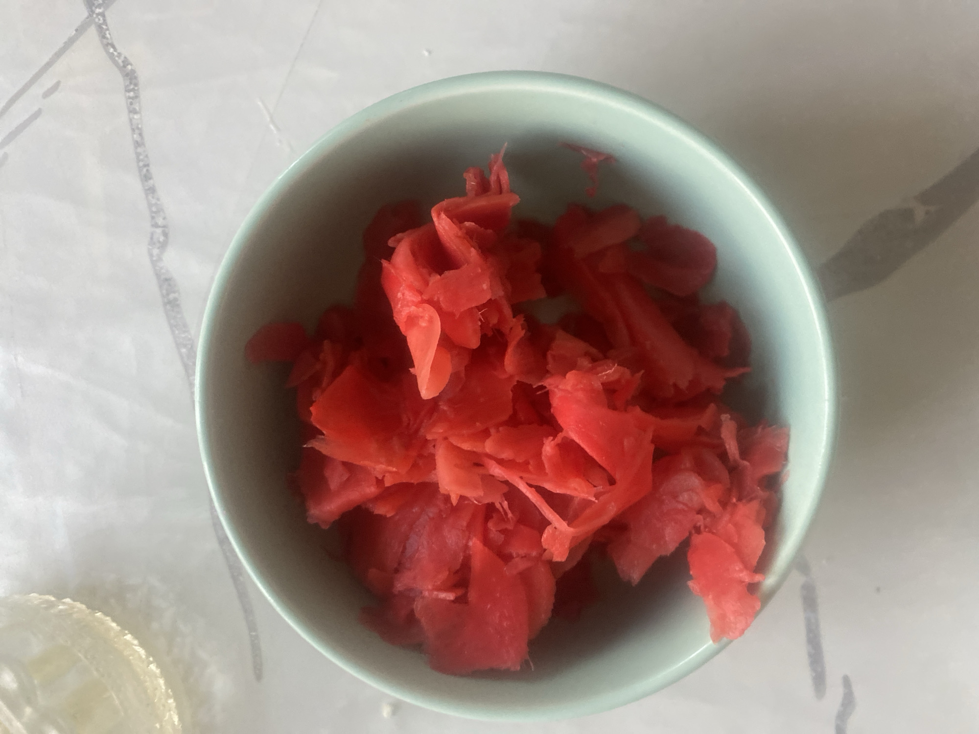 A close-up, overhead shot shows a light teal bowl filled with bright pink pickled ginger. The bowl is situated on a light-colored, textured surface that appears to be a countertop or table. The surface has faint, dark gray, irregular lines running across it, suggesting a pattern or texture. To the upper left of the bowl, a portion of a clear glass with a light yellow liquid is visible, its rim reflecting light. The overall lighting is bright, with some shadows visible on the surface, particularly a elongated shadow extending from the bowl. The image is located in the city of Boraldai, Kazakhstan.