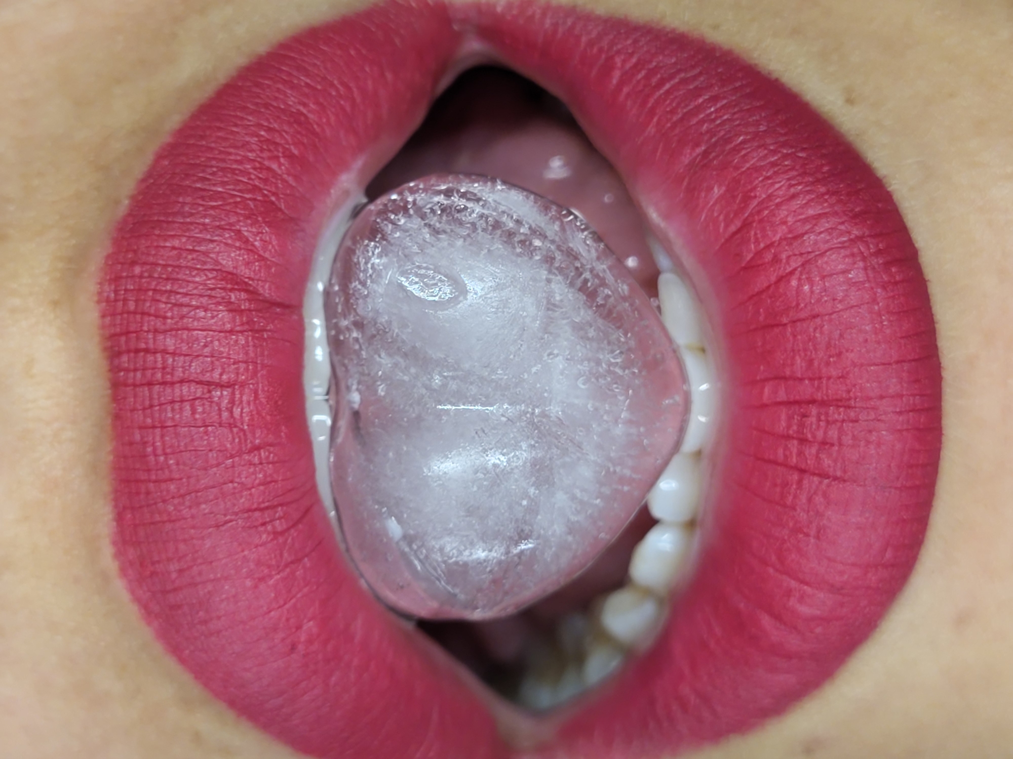 A close-up, eye-level, indoor shot shows a person's open mouth, with a heart-shaped ice cube on their tongue. The lips are painted with matte, raspberry-pink lipstick. The ice cube is translucent and partially melted, with small bubbles trapped inside. The teeth are visible and appear white and healthy. The skin surrounding the mouth is light-toned with visible pores and some freckles. The background is out of focus. This image was captured in Horishni Plavni, Ukraine.