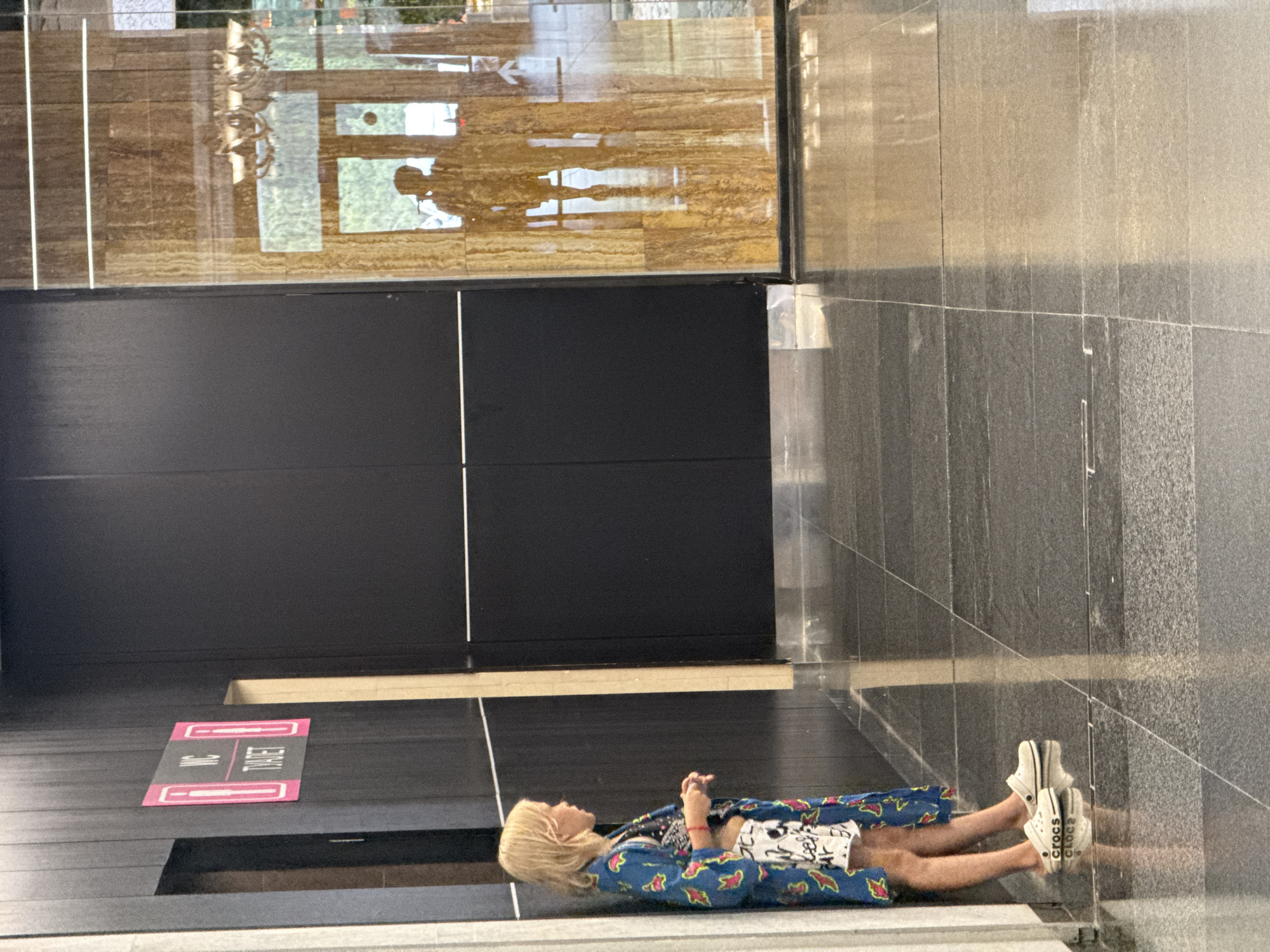 A young girl with blonde hair stands leaning against a dark wall. She is wearing a blue and white patterned robe over a black and white graphic top and white shorts. She is also wearing white Crocs. The wall behind her has a sign with "WC" and figures indicating men's and women's restrooms. To her right, a glass wall with a wood-grain pattern reflects a chandelier and the silhouette of a person. The floor is tiled with dark grey and black polished stone. The scene appears to be an indoor public space, possibly a mall or hotel lobby.