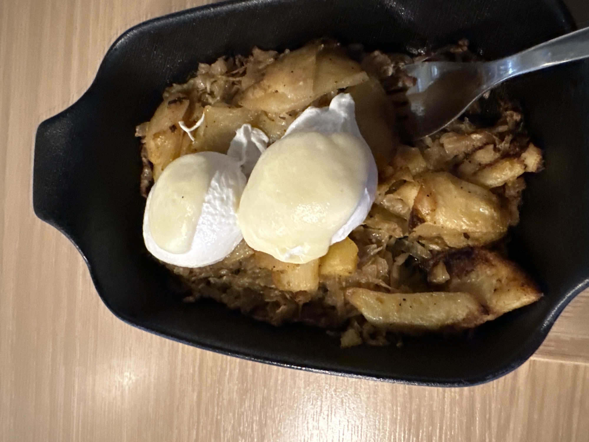 An overhead shot captures a dish served in a black, possibly cast iron, rectangular bowl with small handles. The main components of the meal are golden-brown, pan-fried or roasted potato wedges, mixed with what appears to be sautéed onions or finely chopped cabbage, forming a base layer. The potatoes show signs of browning and some darker, caramelized edges.
Two poached eggs are prominently placed on top of the potato and onion mixture. The egg whites are firm and opaque, while the yolks, visible in varying degrees, are a pale, creamy yellow, suggesting they are soft-cooked. A shiny metal spoon is partially visible entering the dish from the upper right, indicating the meal is either being served or in the process of being eaten.
The serving bowl rests on a light-colored wooden surface, likely a table, with a visible wood grain pattern. The lighting is warm and somewhat dim, possibly artificial, highlighting the textures and colors of the food. There are no people visible, and no text is discernible in the image. The scene suggests a casual dining setting for a breakfast, brunch, or simple meal.