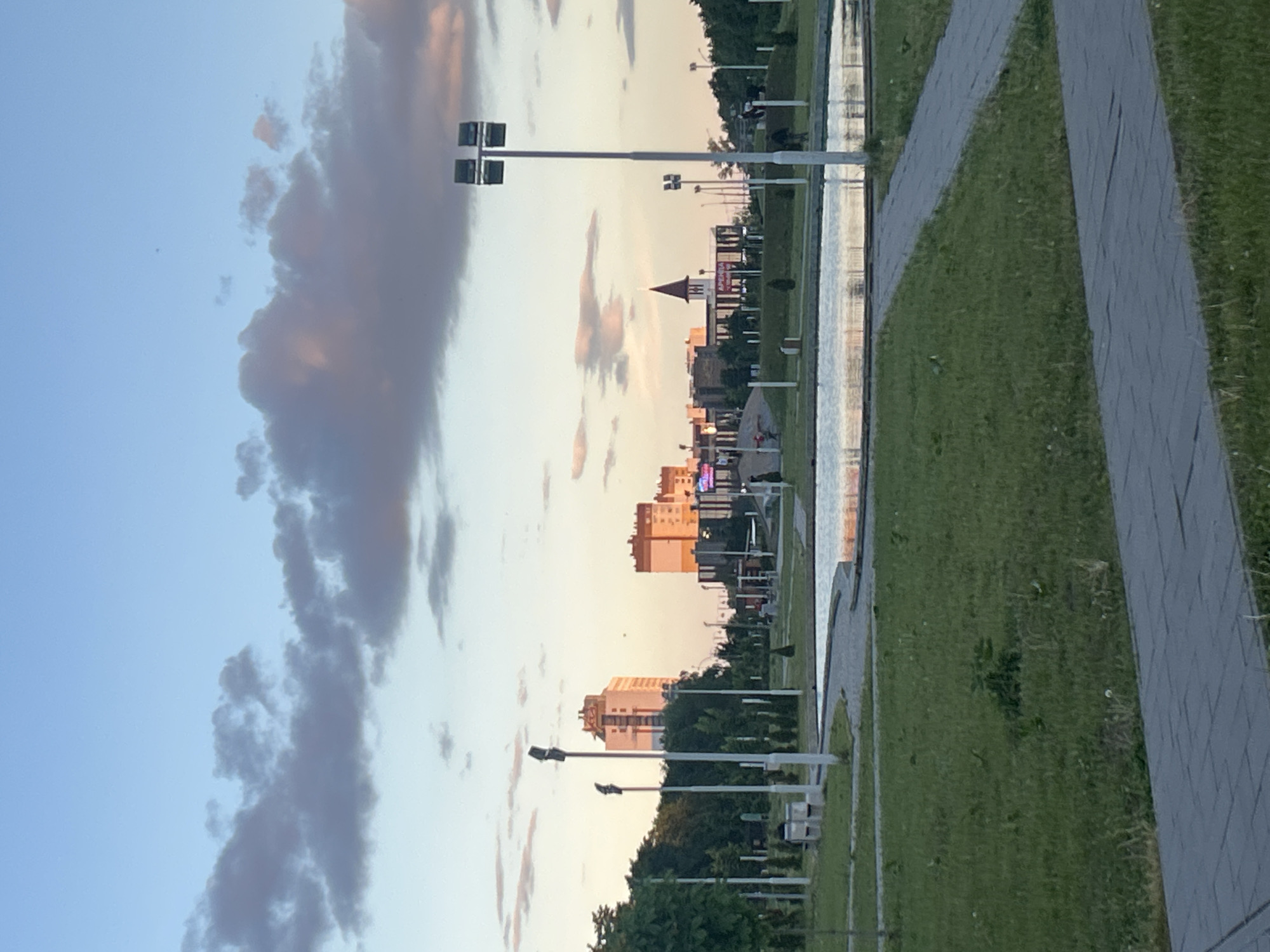 The image shows a cityscape in Tolyatti, Russia, taken during sunset or sunrise. The sky is a mix of light blue and shades of pink and orange. A line of buildings of varying heights is visible in the center. There is a body of water reflecting the buildings and sky, and a paved pathway alongside a grassy area. Light posts are situated along the pathway. Clouds are present in the sky.