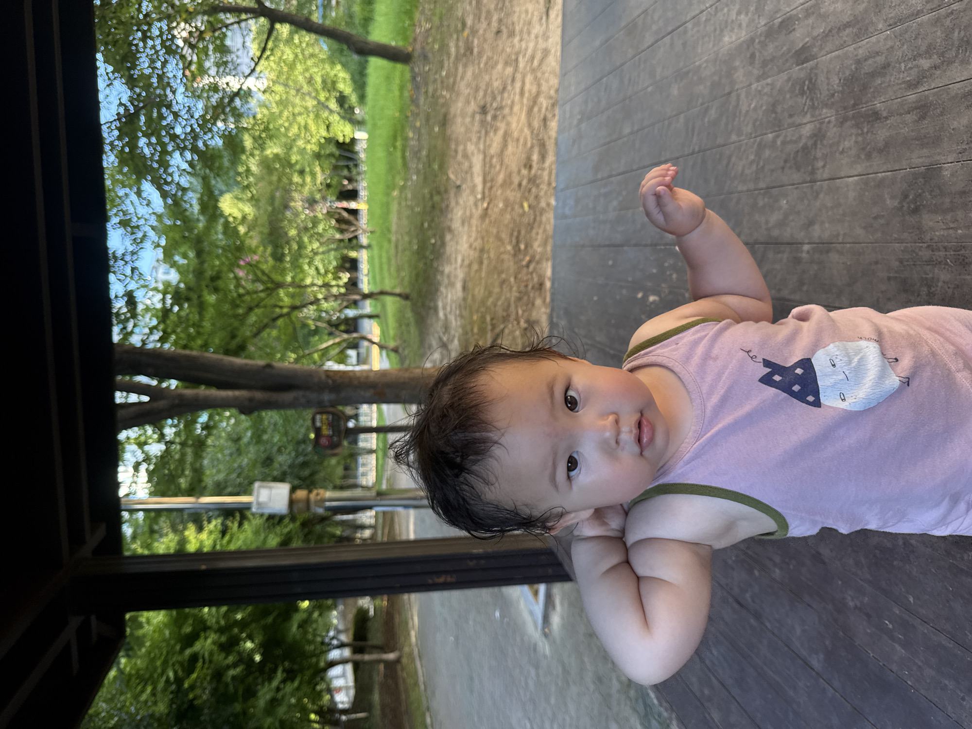 A young child stands on a wooden platform, looking towards the camera. The child is wearing a pink tank top with a graphic print of a cartoon character. The background consists of a park with trees, green grass, and a pathway. A wooden structure with a dark roof is visible to the left. The scene appears to be outdoors, possibly in a public park or recreational area. The lighting suggests it is daytime, with sunlight filtering through the trees.