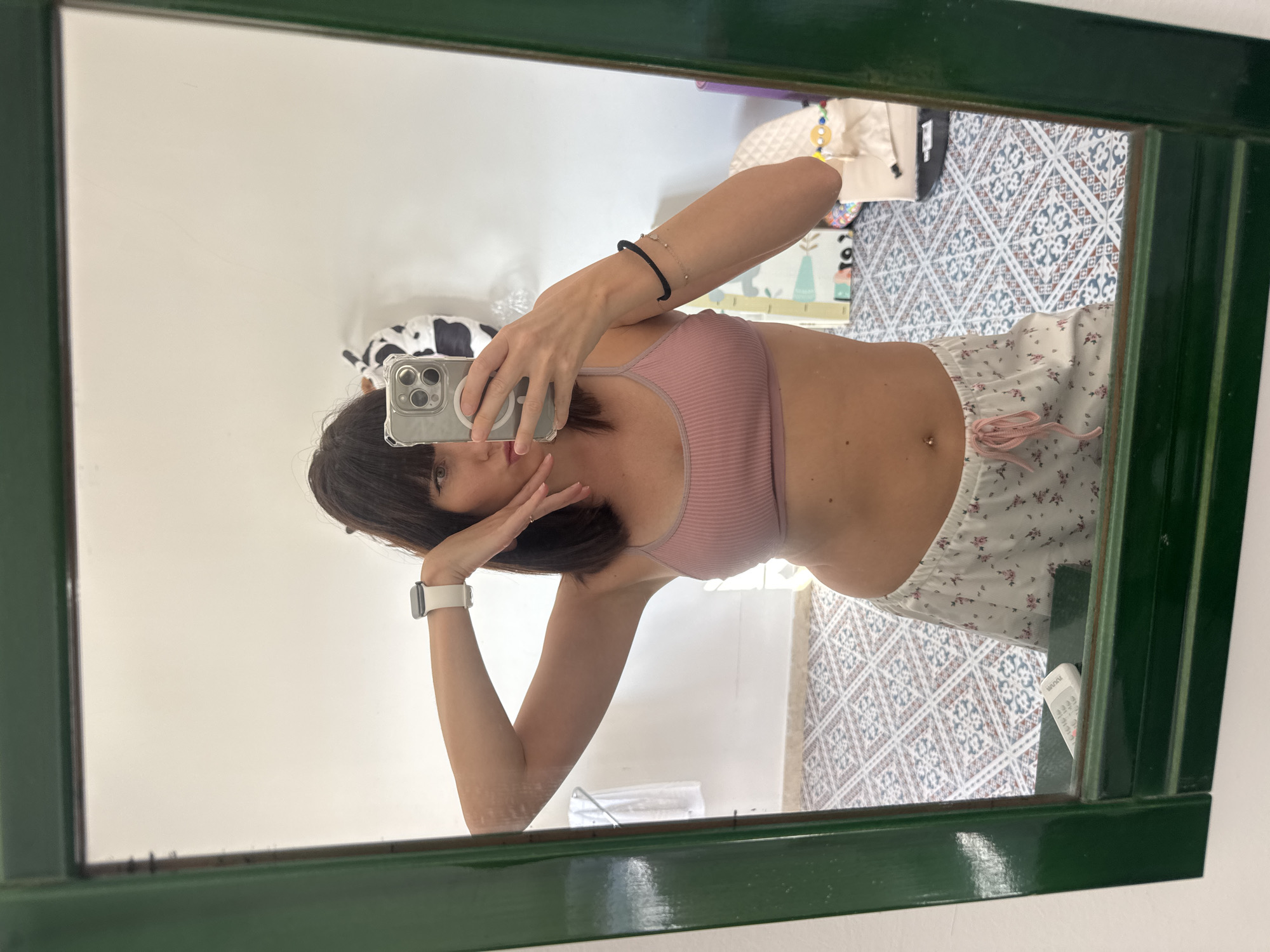 A young woman with dark hair stands in front of a mirror in what appears to be a residential bathroom. She is wearing a pink bralette and white pajama shorts with a floral pattern. She holds a smartphone in her right hand, taking a selfie. Her left arm is raised, and her hand is near her face. The mirror reflects a white wall, a floor with a patterned tile, and some objects in the background, including a stuffed cow toy, a baby mat, and a remote control on the mirror's ledge. The bathroom has a green-framed mirror and a white wall. The environment suggests a domestic setting.