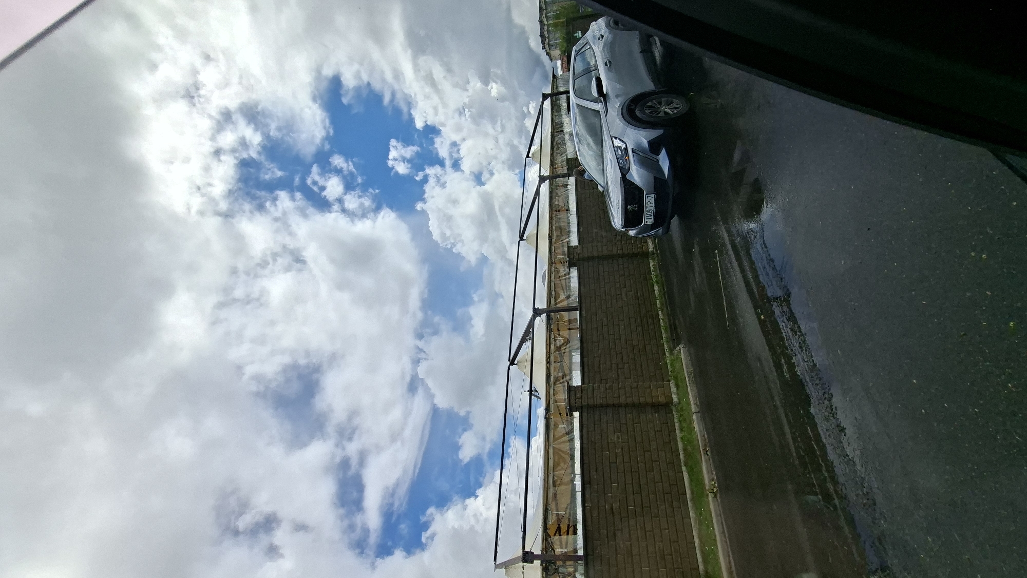 In Myadzyel, Belarus, on a wet asphalt road, a silver Peugeot 2008 SUV is parked. The vehicle's license plate reads "1409 TP-7". In the background, a brick wall runs along the side of the road, and behind it, a long, covered structure with white canopies is visible. The sky is filled with dramatic, dark clouds interspersed with patches of blue. The scene appears to be on a cloudy day, possibly after rain.